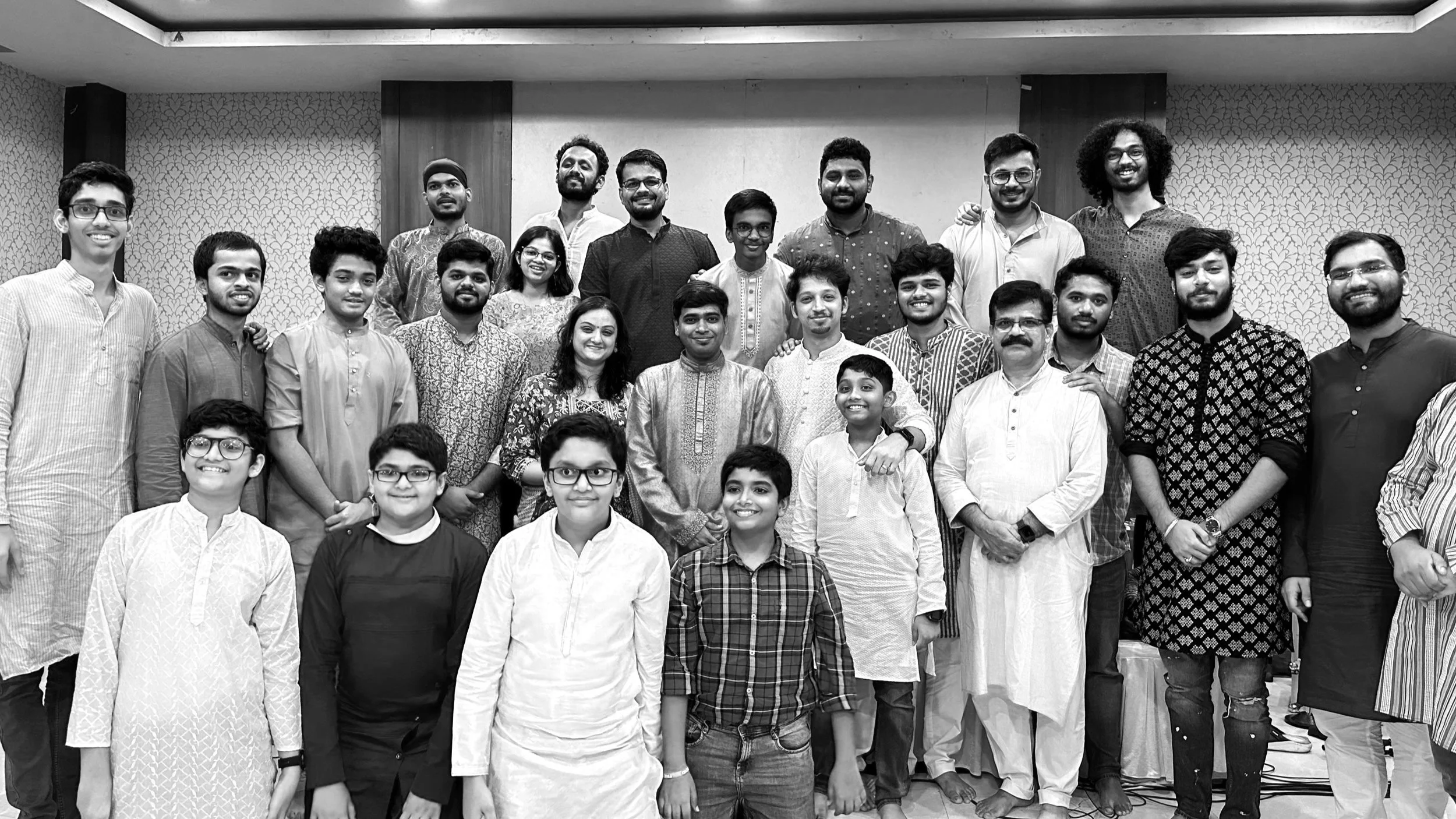 Group portrait of around 23 people, mostly young and middle-aged, posing together in a room with patterned wallpaper, some wearing traditional South Asian clothing, smiling at the camera.