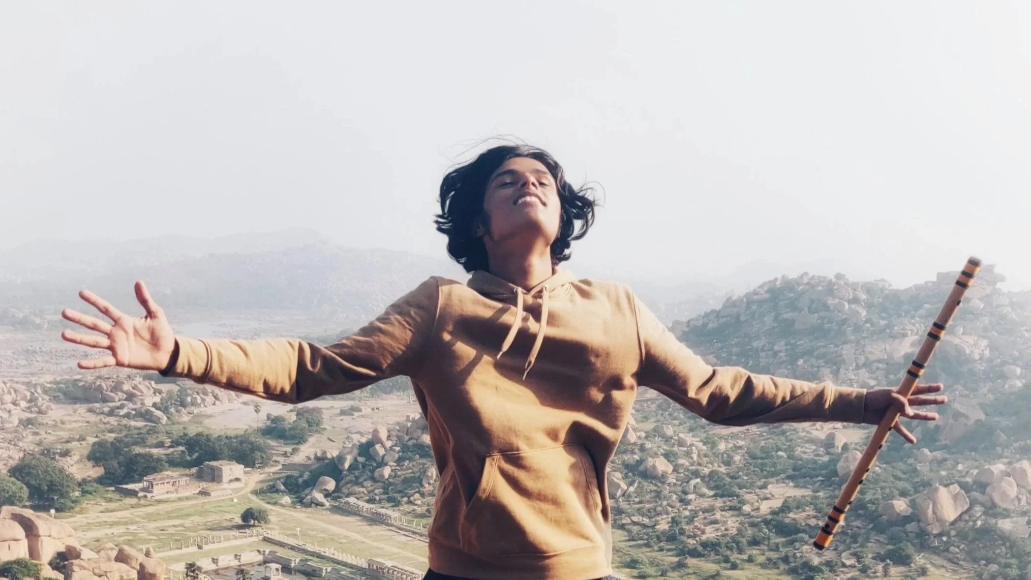 Person standing with arms outstretched on a hilltop, holding a flute, with rocky hills and a valley in the background.