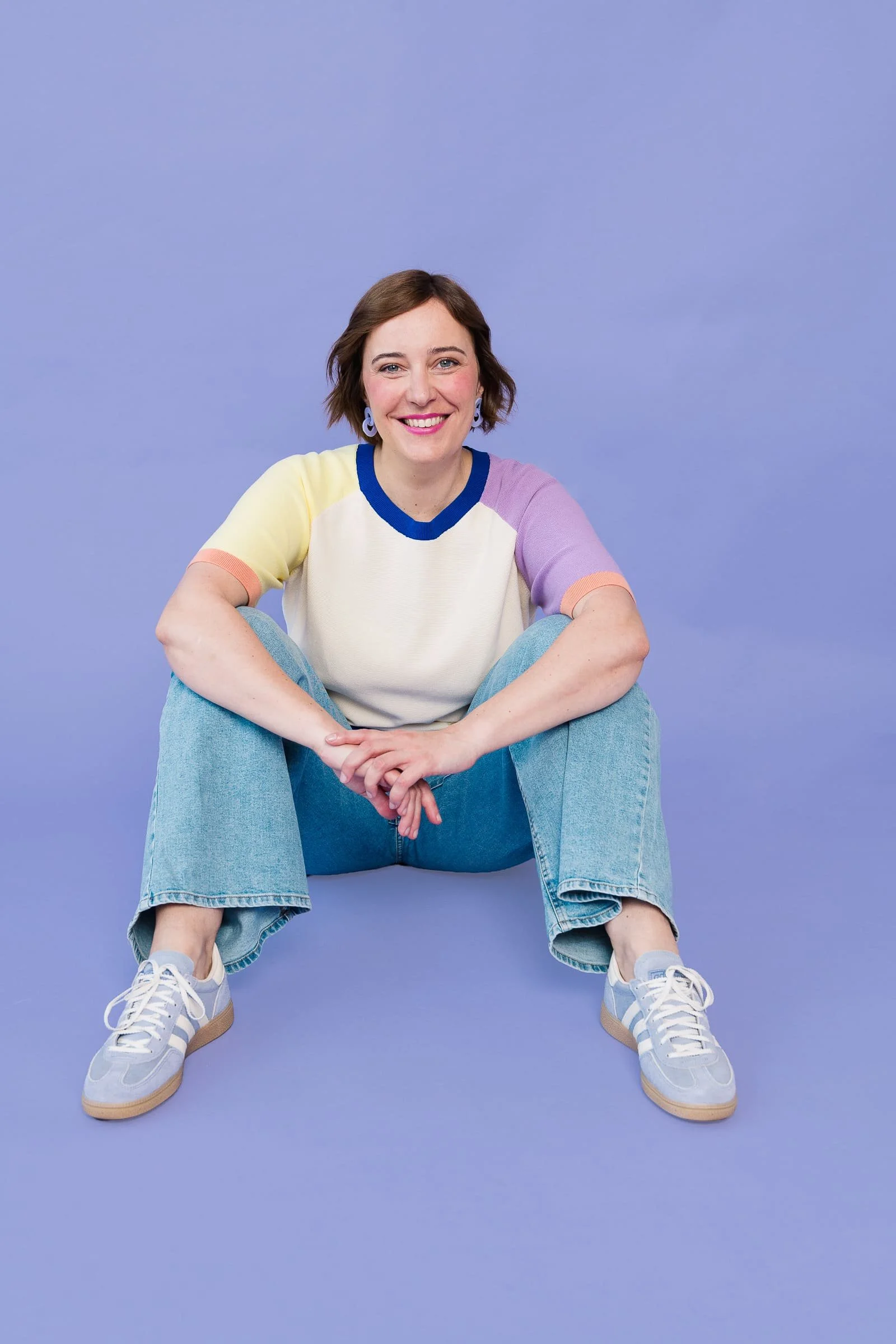 Portrait von Ina Kimmel | THERAPIEPAUSE®. Sie sitzt auf dem Boden vor einem lilafarbenen Hintergrund. Die Arme liegen auf ihren Knien. Sie lacht in die Kamera.