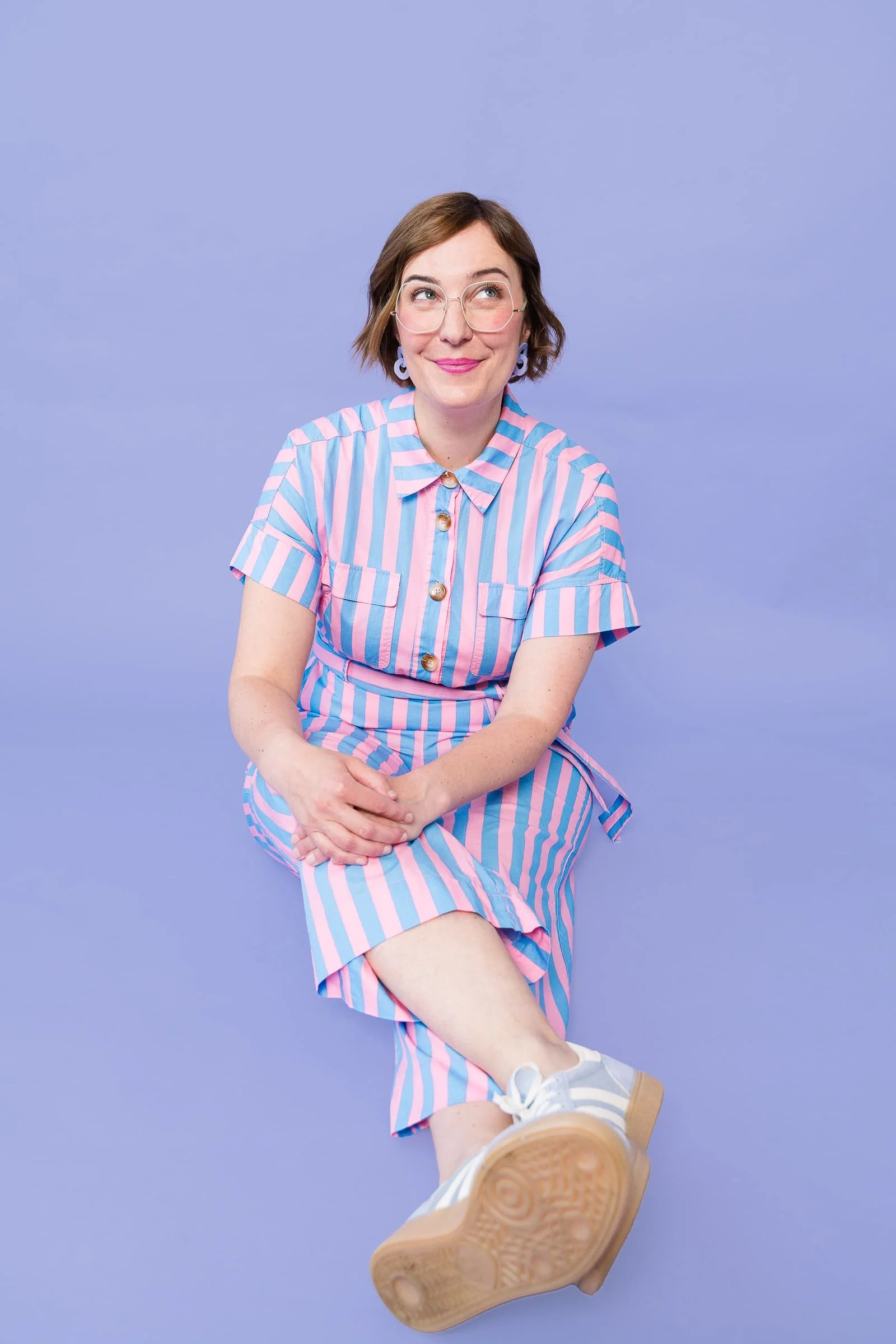 Portrait von Ina Kimmel, Gründerin und Coach bei THERAPIEPAUSE®, sie sitzt auf dem Boden auf einem lilafarbenen Hintergrund, trägt einen bunt-gestreiften Jumpsuit und  schaut lächelnd nach oben