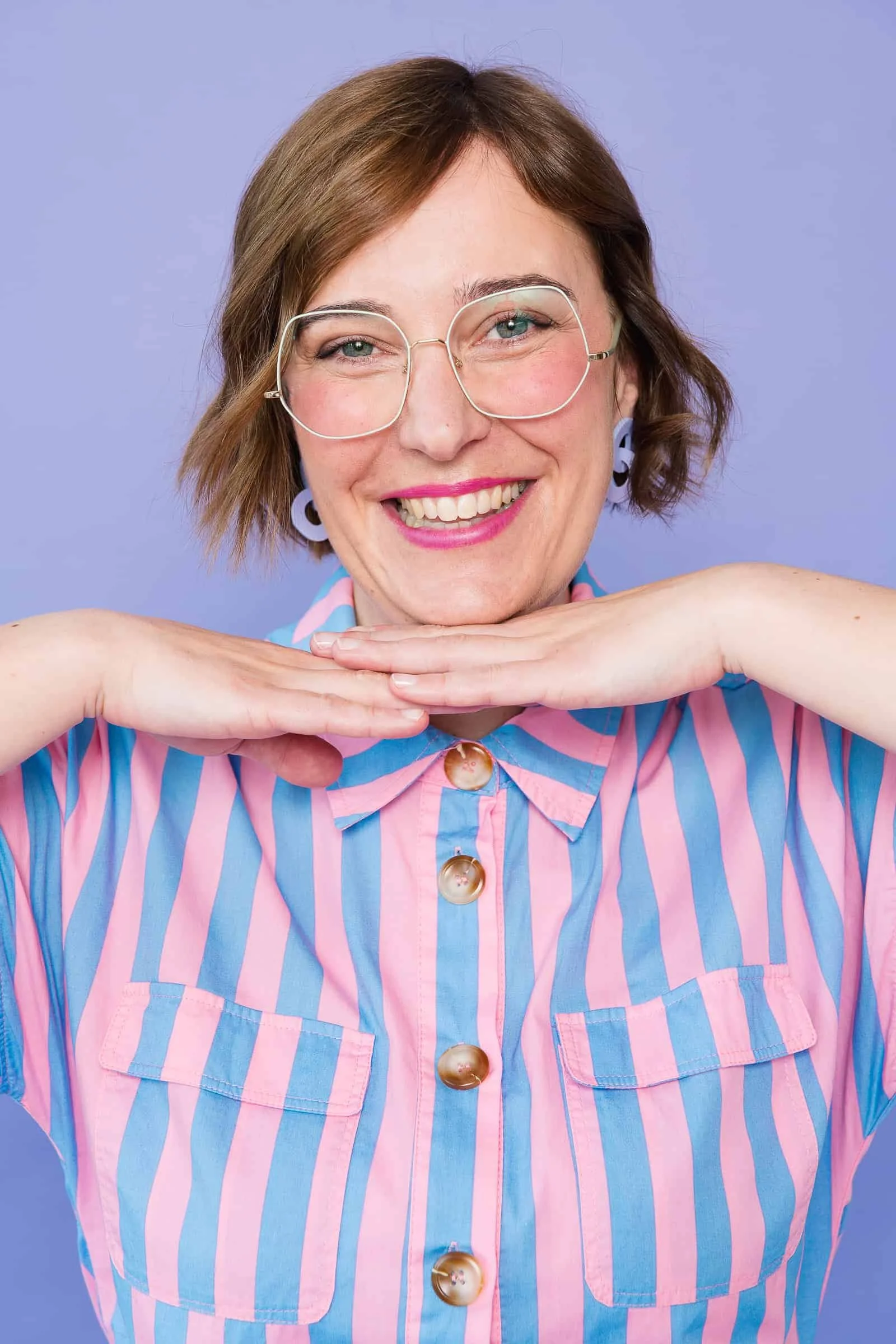 Portrait von Ina Kimmel | THERAPIEPAUSE®. Ihre Hände liegen unter ihrem Kinn. Sie lächelt in die Kamera. Sie trägt eine große Brille und lilafarbene Ohrringe.