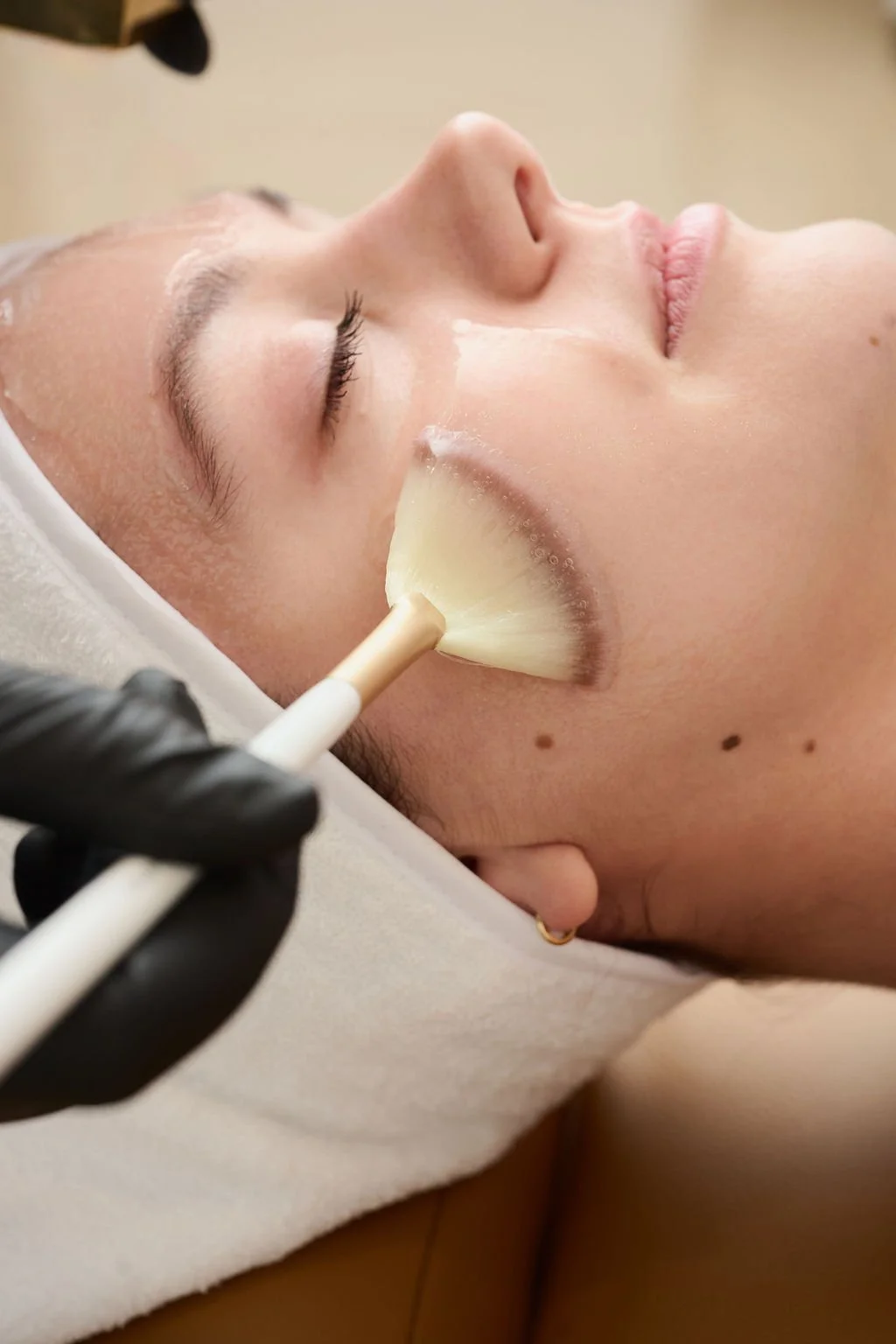 A person wearing black gloves holding a brush with foam in a small bowl near a person's face, suggesting a skincare or facial treatment in progress.