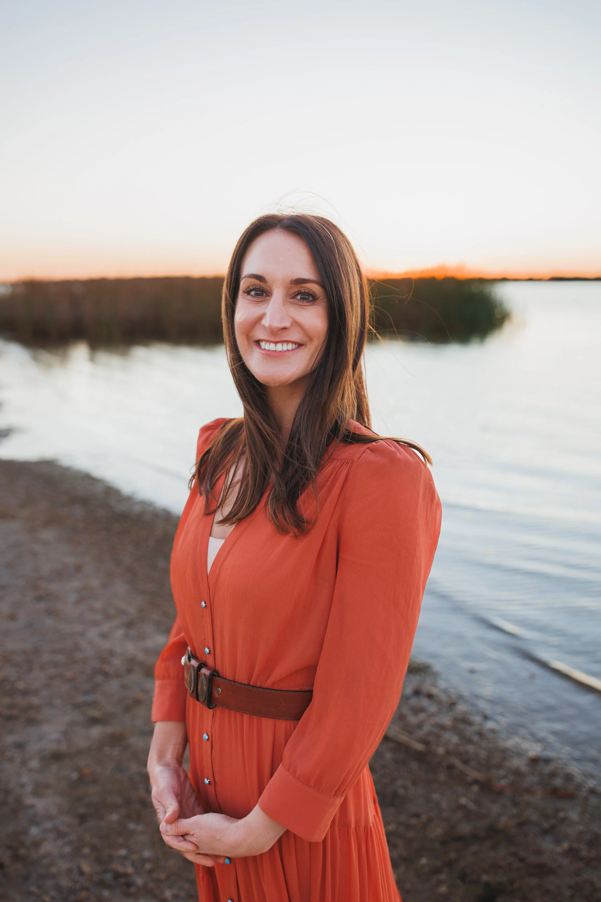 A smiling woman with long brown hair wearing an orange dress standing by a lake at sunset.