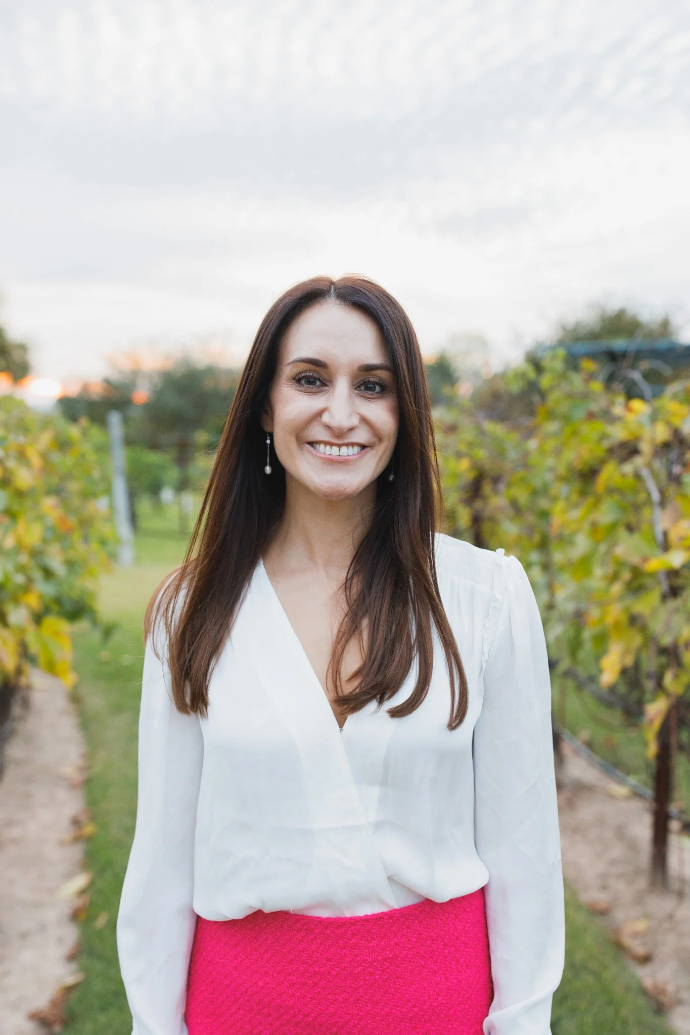 A woman with long brown hair and pearl earrings standing outdoors in a vineyard, smiling at the camera, wearing a white blouse and pink skirt, with green foliage and cloudy sky in the background.