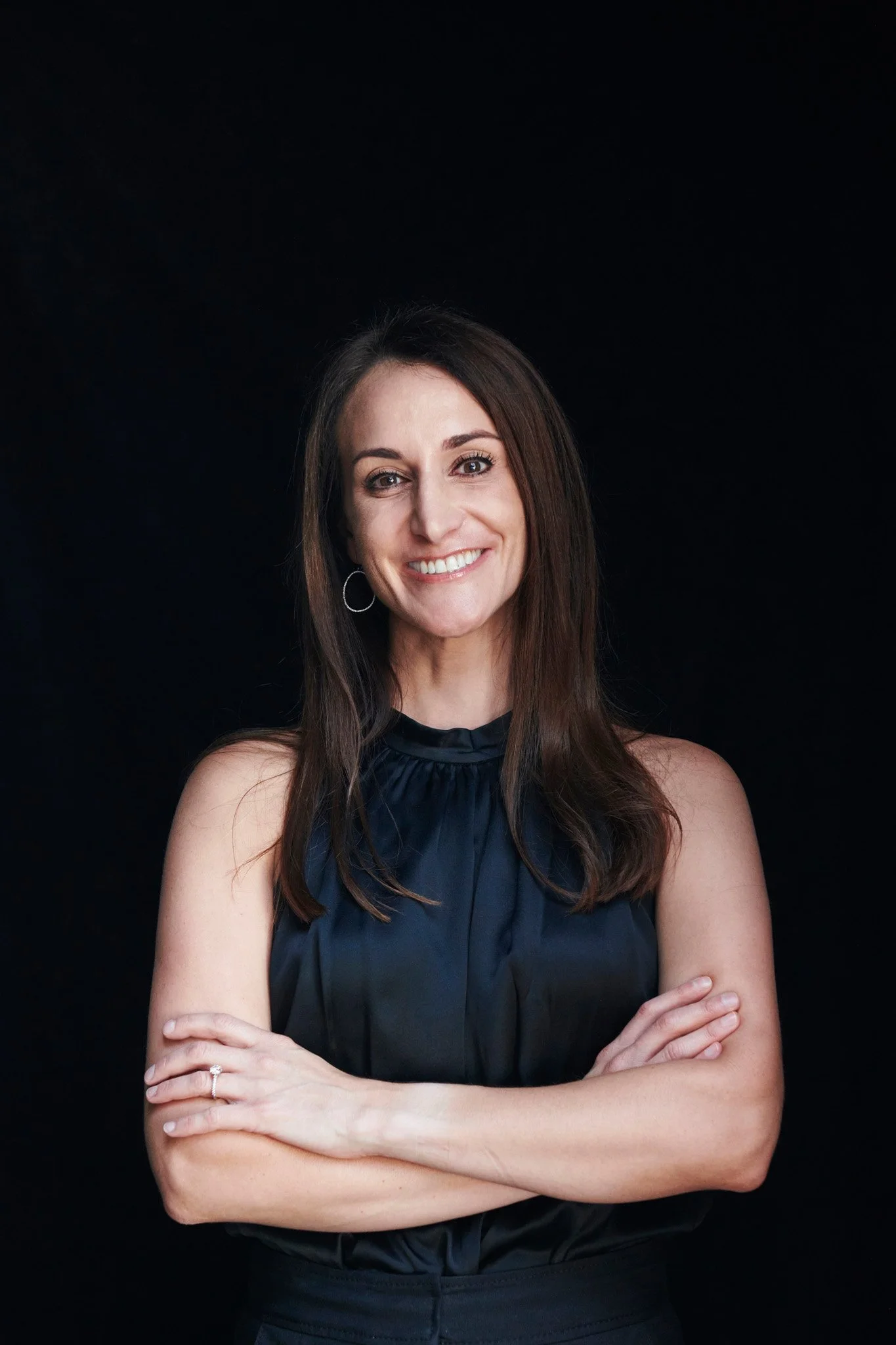 Smiling woman with crossed arms, wearing a sleeveless black top, against a black background.