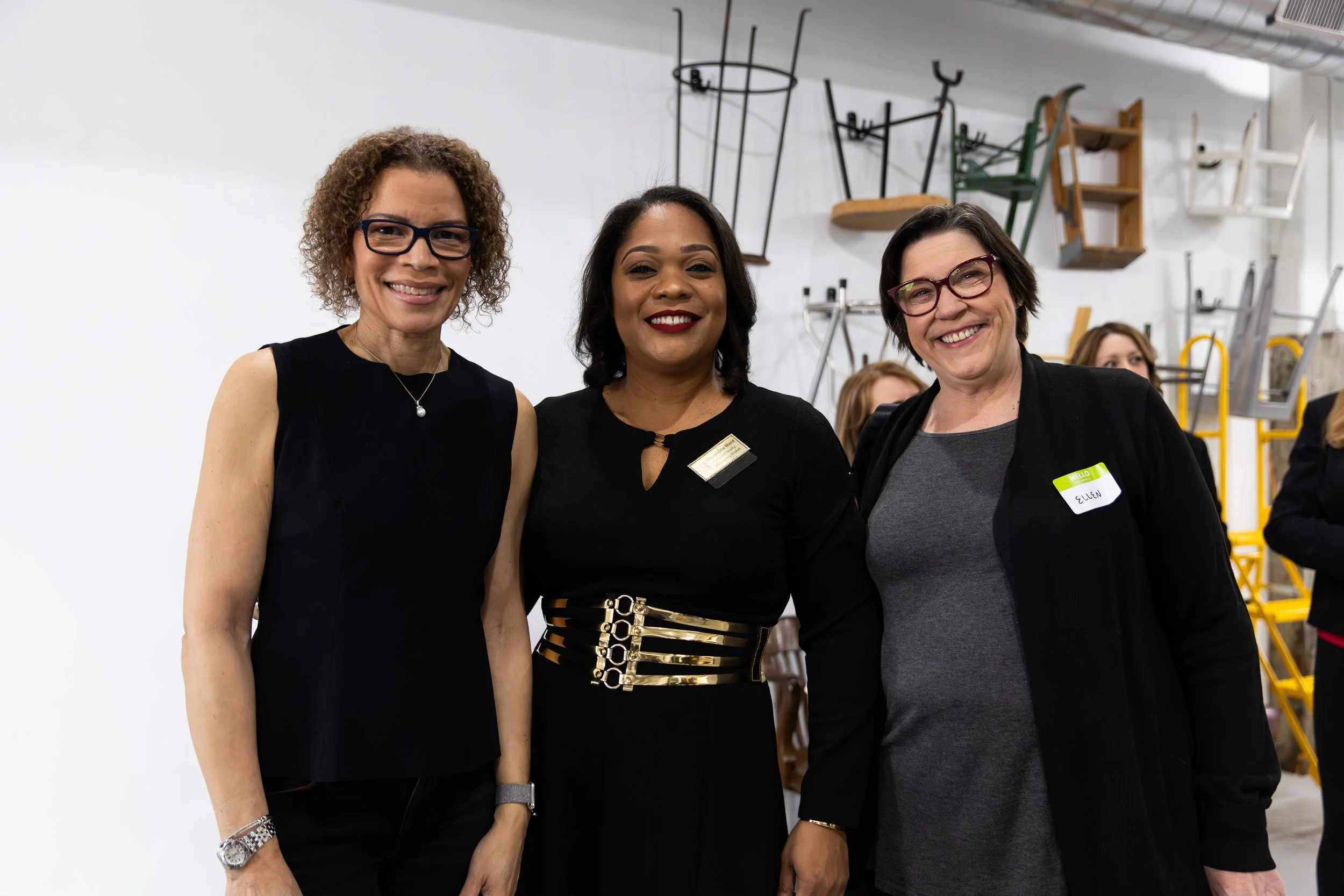 Three women stand side by side, smiling, with chairs hanging on the wall behind them in a well-lit room.