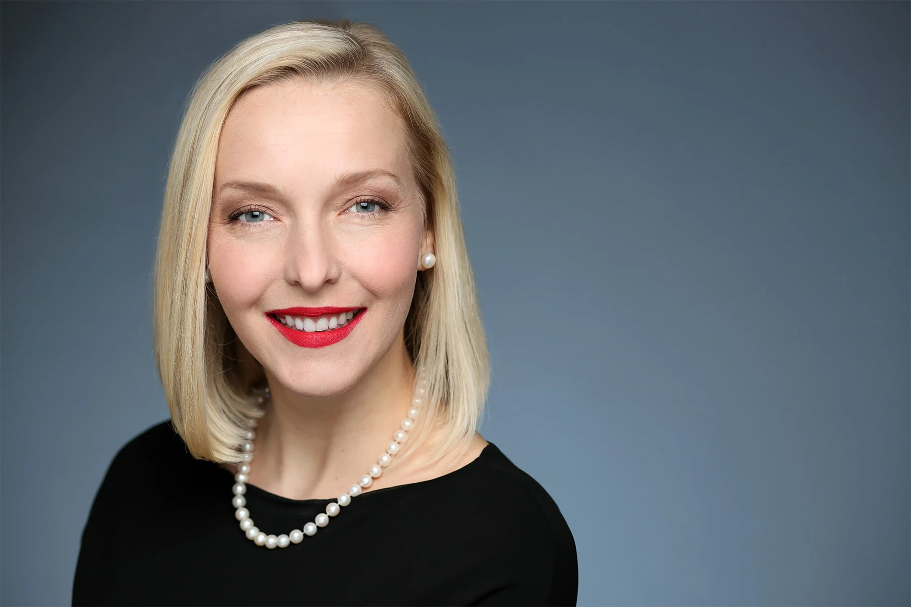 A blonde woman with light skin, wearing red lipstick, a pearl necklace, pearl earrings, and a black top, smiling against a gray background.