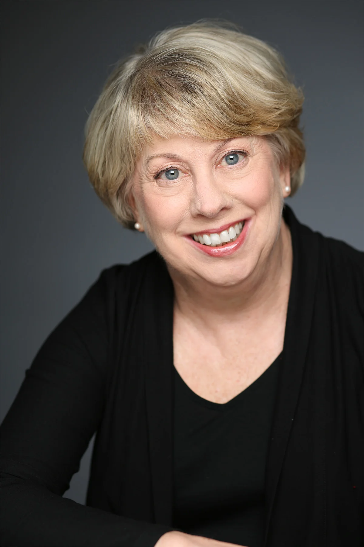 Portrait of a smiling middle-aged woman with short blonde hair, blue eyes, wearing a black top, and pearl earrings against a dark gray background.