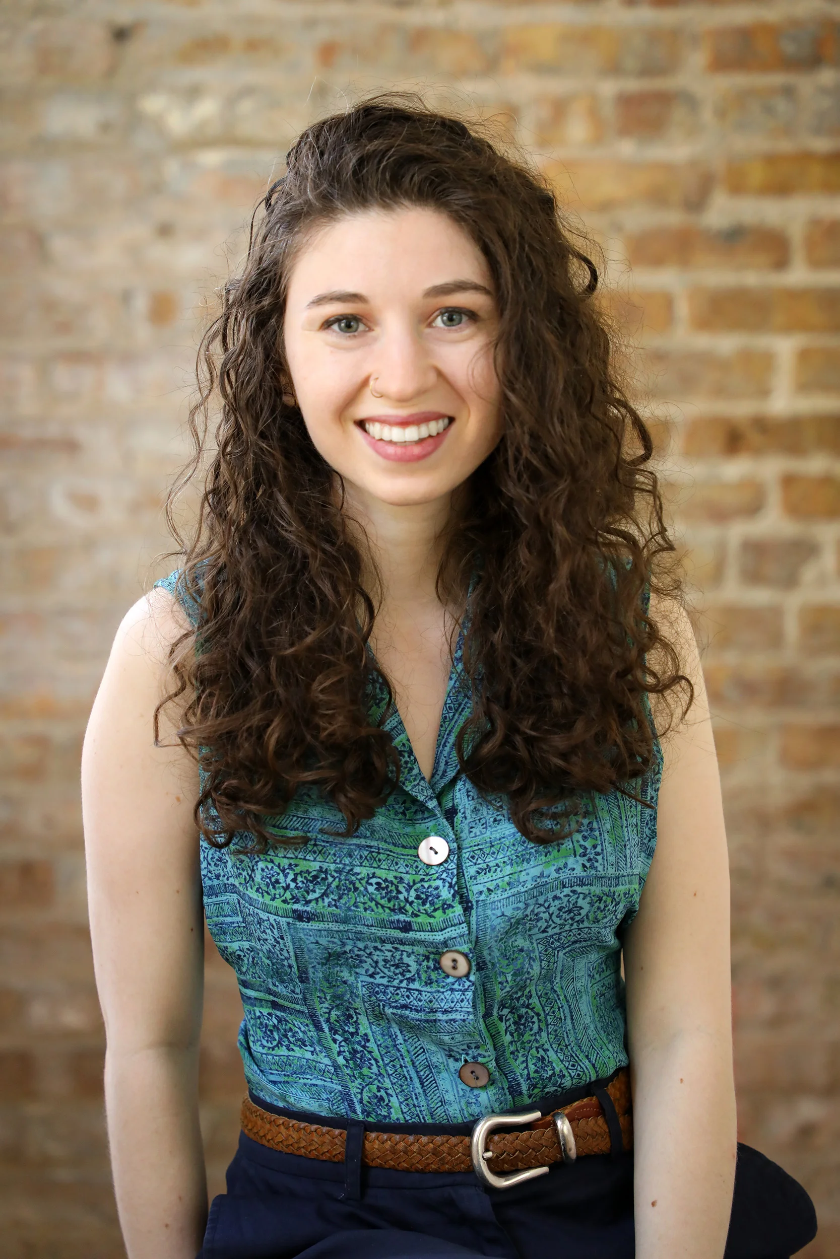 A young woman with curly brown hair and green eyes smiling, wearing a blue patterned sleeveless top and a brown woven belt, sitting against a brick wall background.