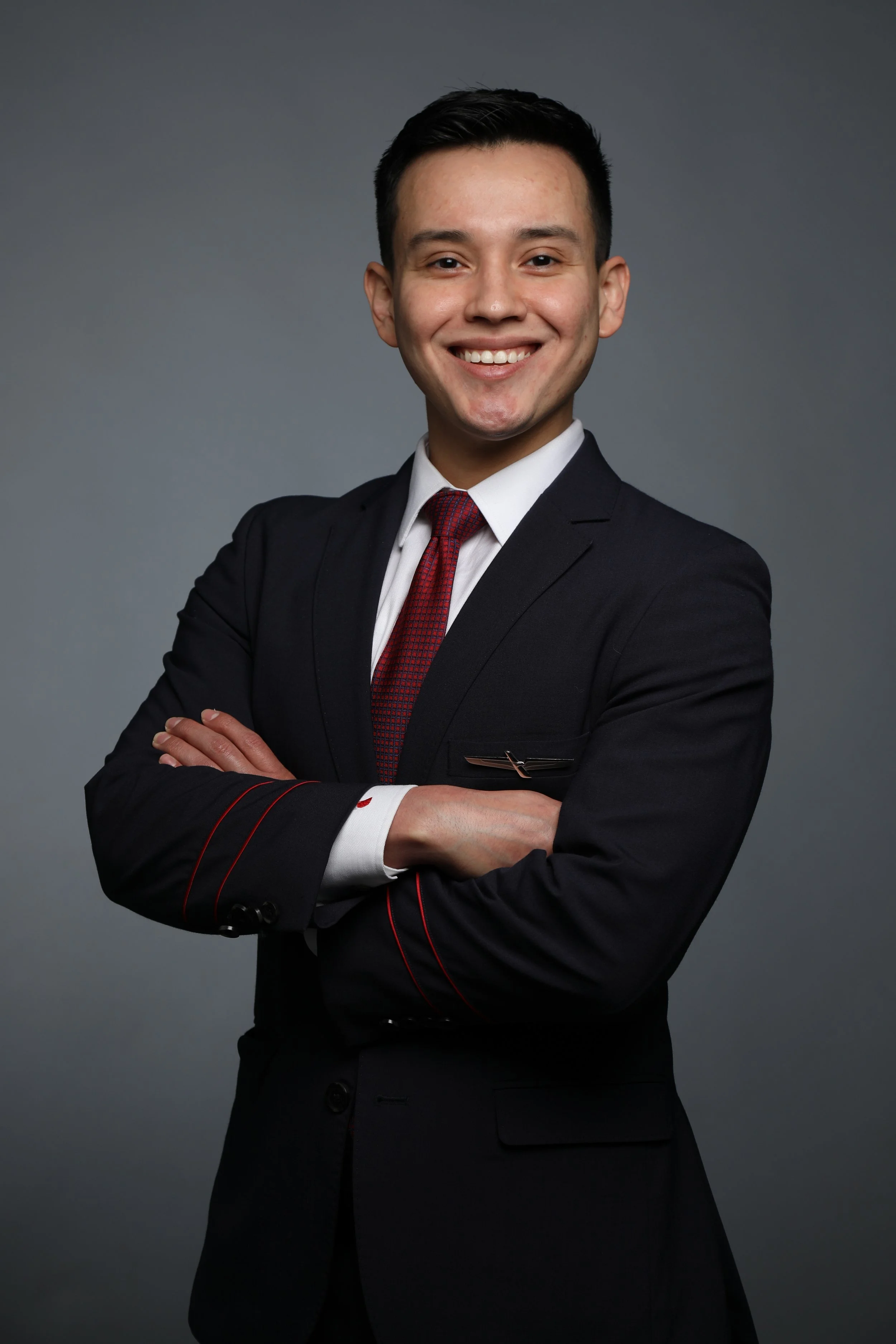 Portrait of a young man in a dark suit with a red tie, smiling with crossed arms, against a gray background.