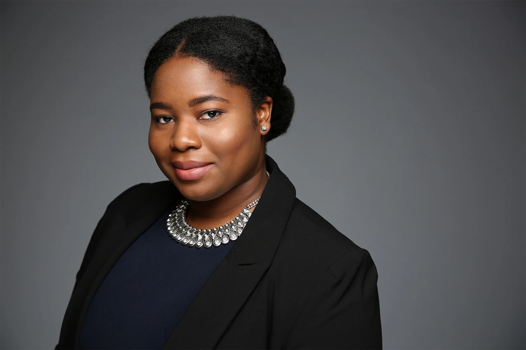 Professional portrait of a Black woman with short curly hair, wearing a black blazer, dark blue top, silver jewelry, and a subtle smile, against a plain gray background.