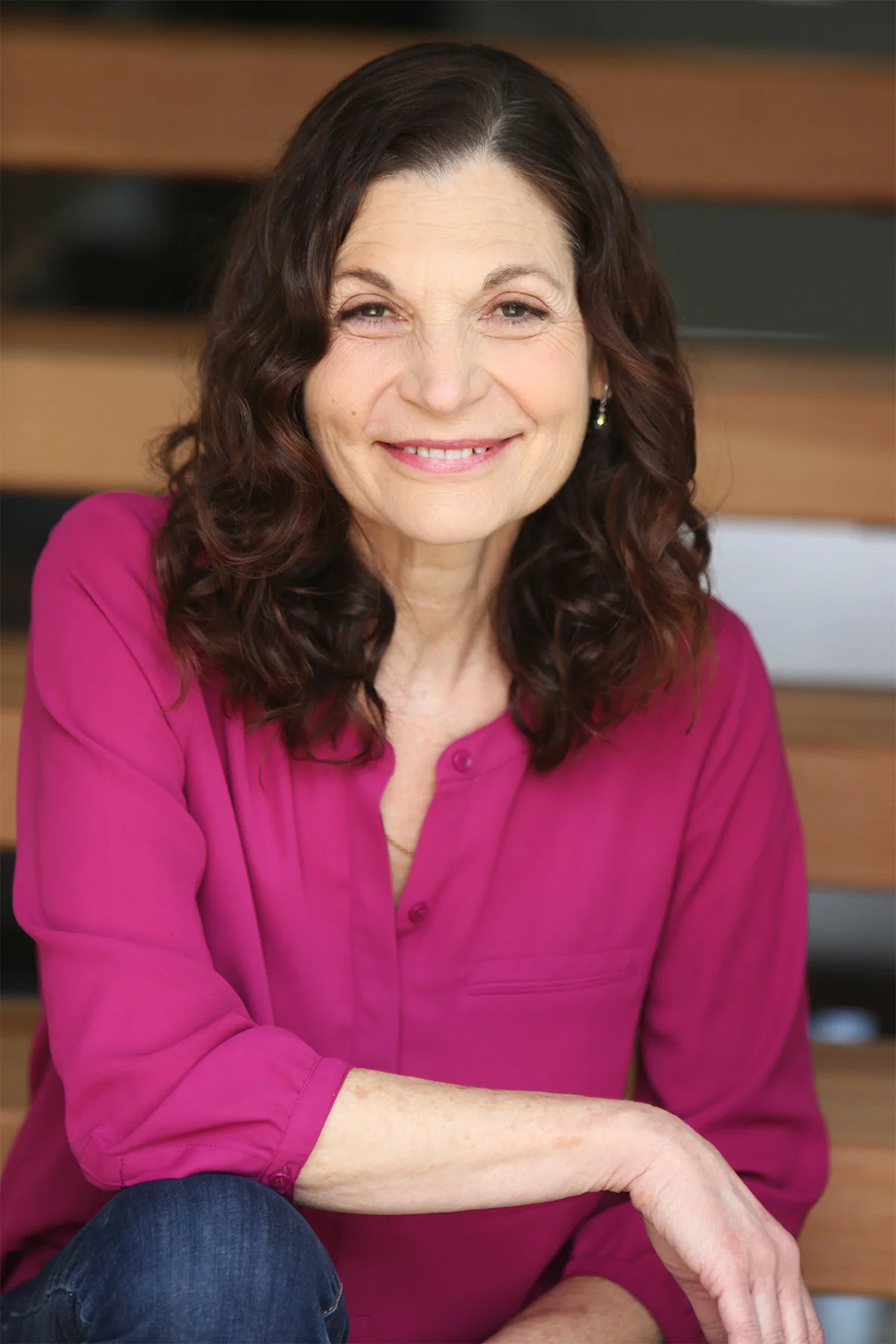 Portrait of a middle-aged woman with dark wavy hair, wearing a bright pink blouse, smiling in front of a wooden background.
