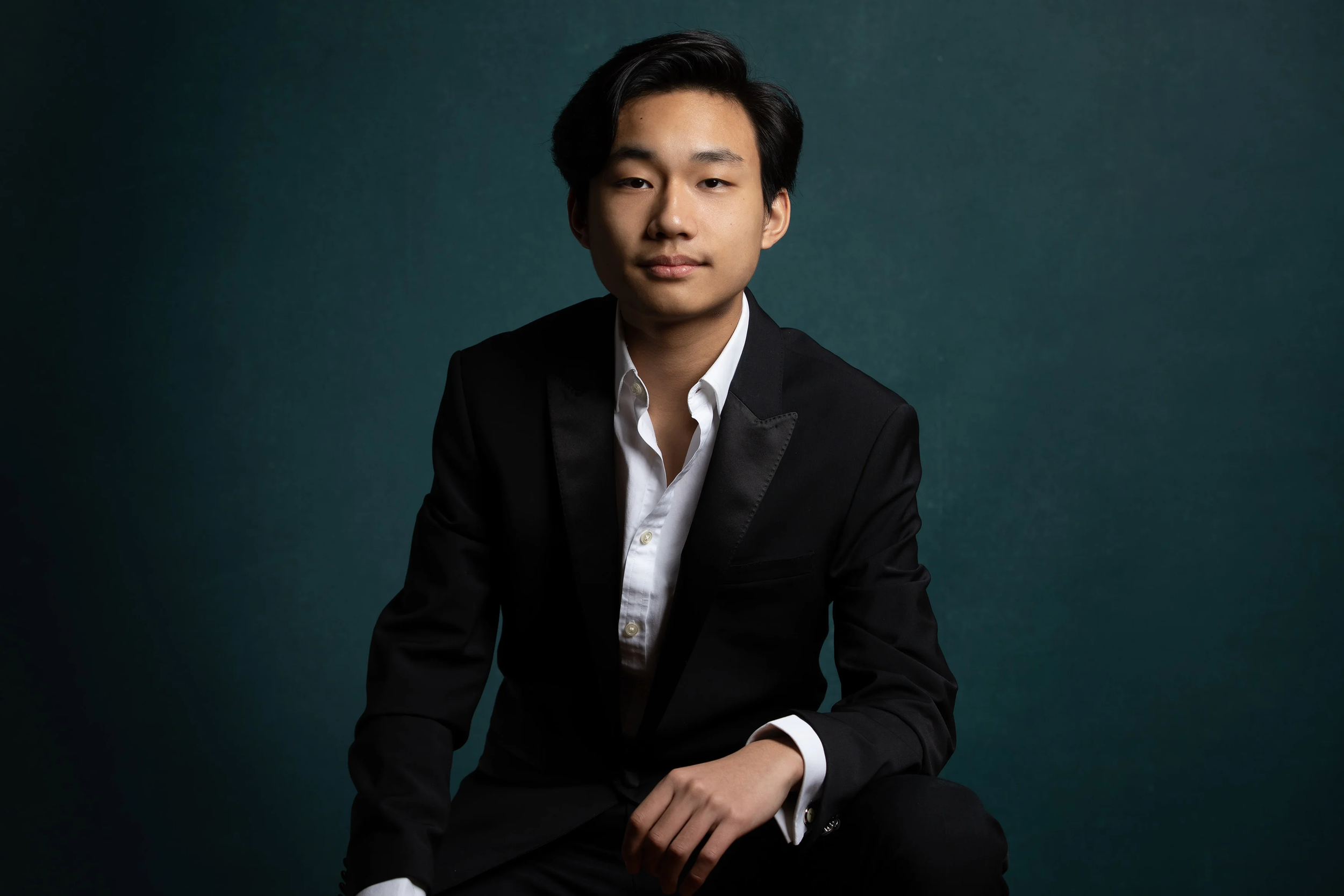 A young man in a black suit with a white shirt, sitting against a dark teal background, looking confidently at the camera.