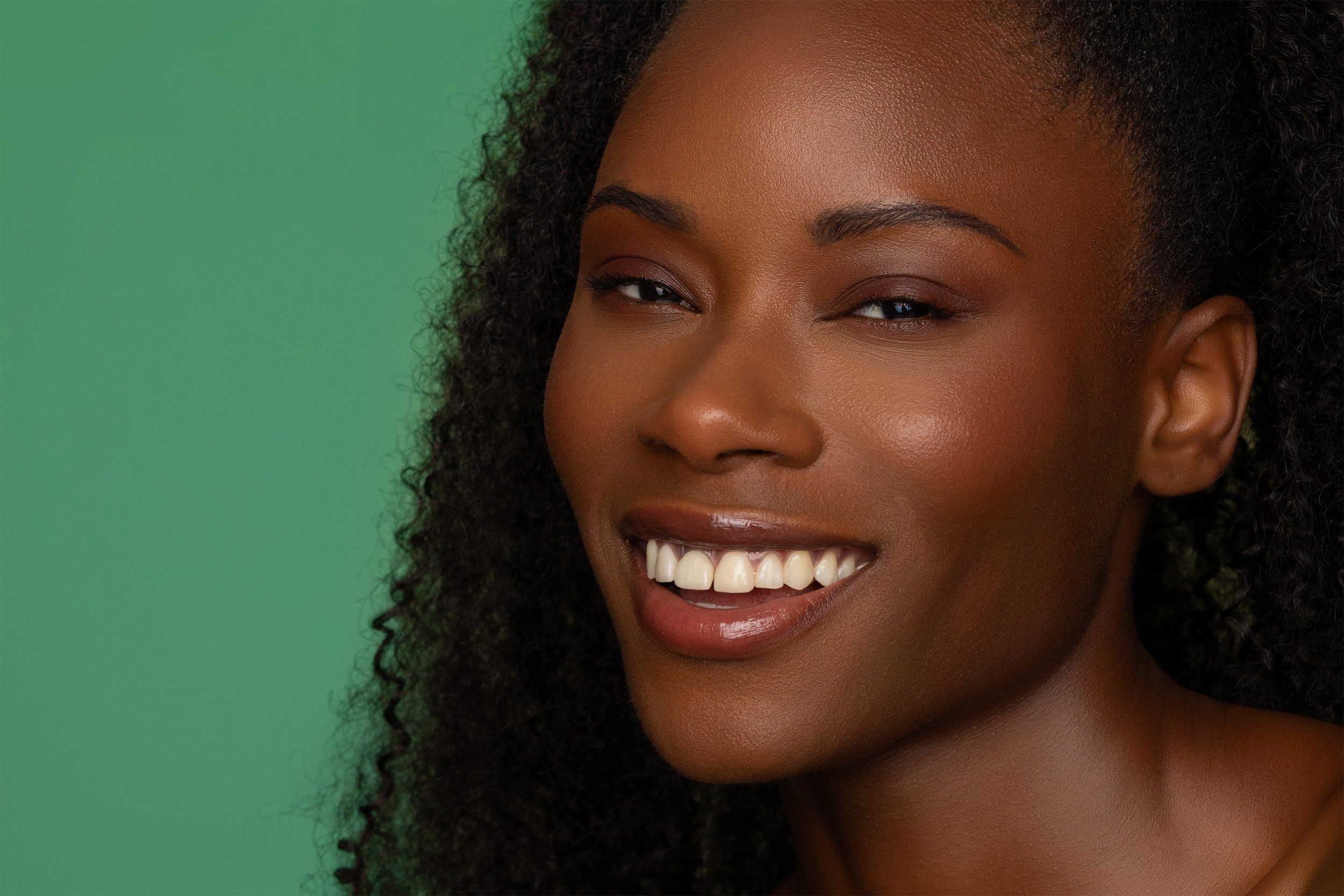 Close-up of a woman with dark skin, curly hair, smiling, against a green background.