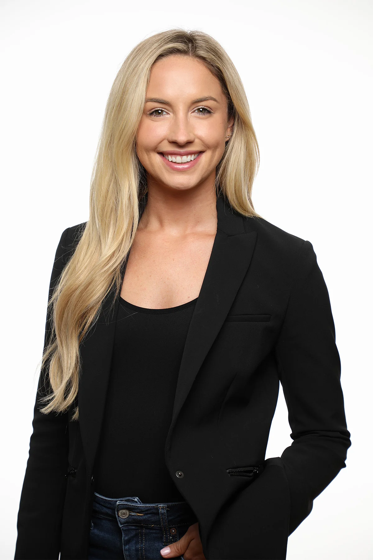 A smiling woman with long blonde hair, wearing a black blazer over a black top and blue jeans, standing against a white background.