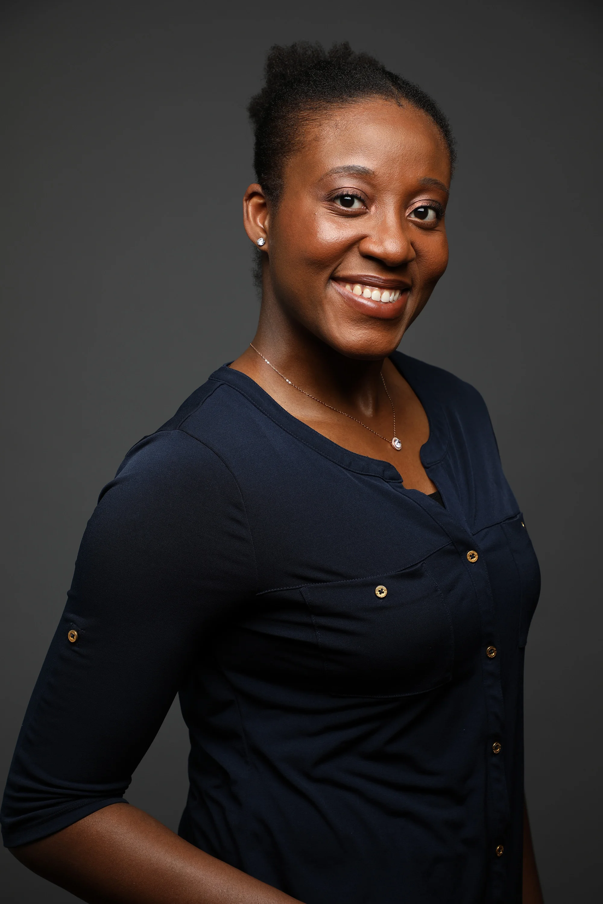 Portrait of a smiling African American woman with natural hair, wearing a dark navy top, stud earrings, and a necklace, against a dark gray background.