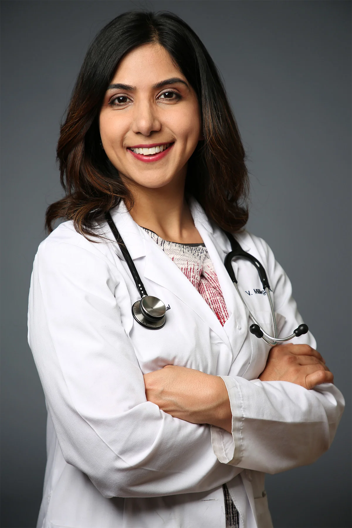 A female doctor with dark hair, wearing a white lab coat and a stethoscope around her neck, smiling and crossing her arms against a gray background.
