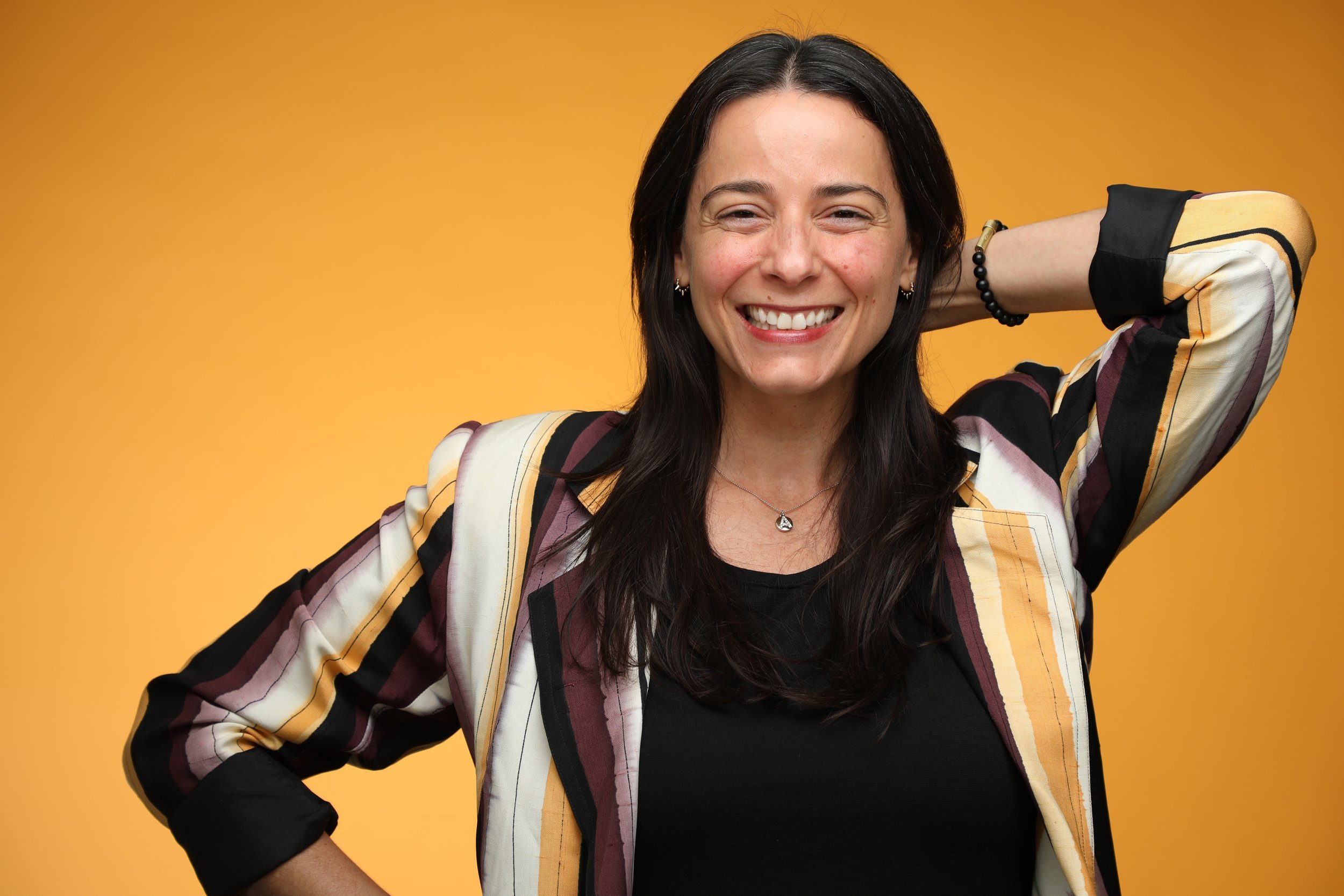 A woman with long dark hair smiling, wearing a patterned blazer over a black top, with her left hand behind her head against a yellow background.