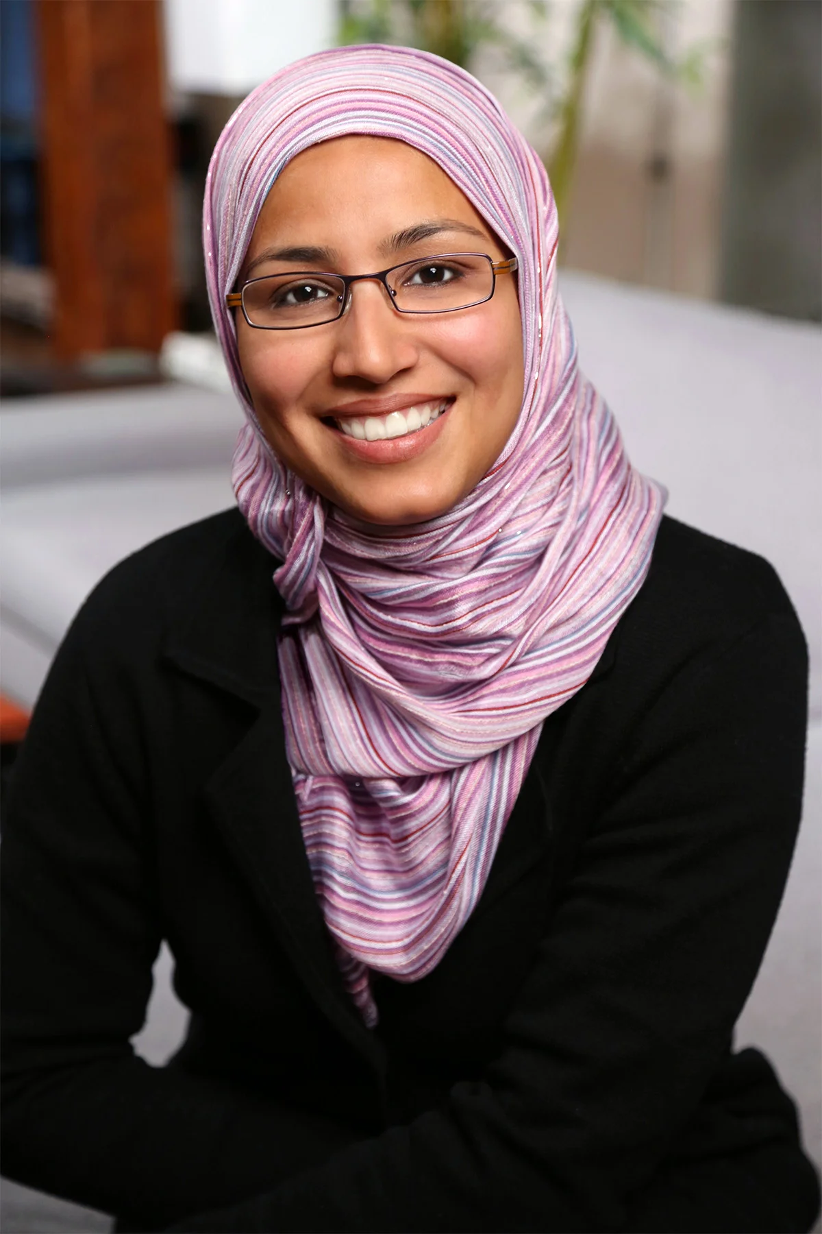 A woman wearing glasses and a pink and white striped hijab smiling at the camera, dressed in a black top, with a blurred background.