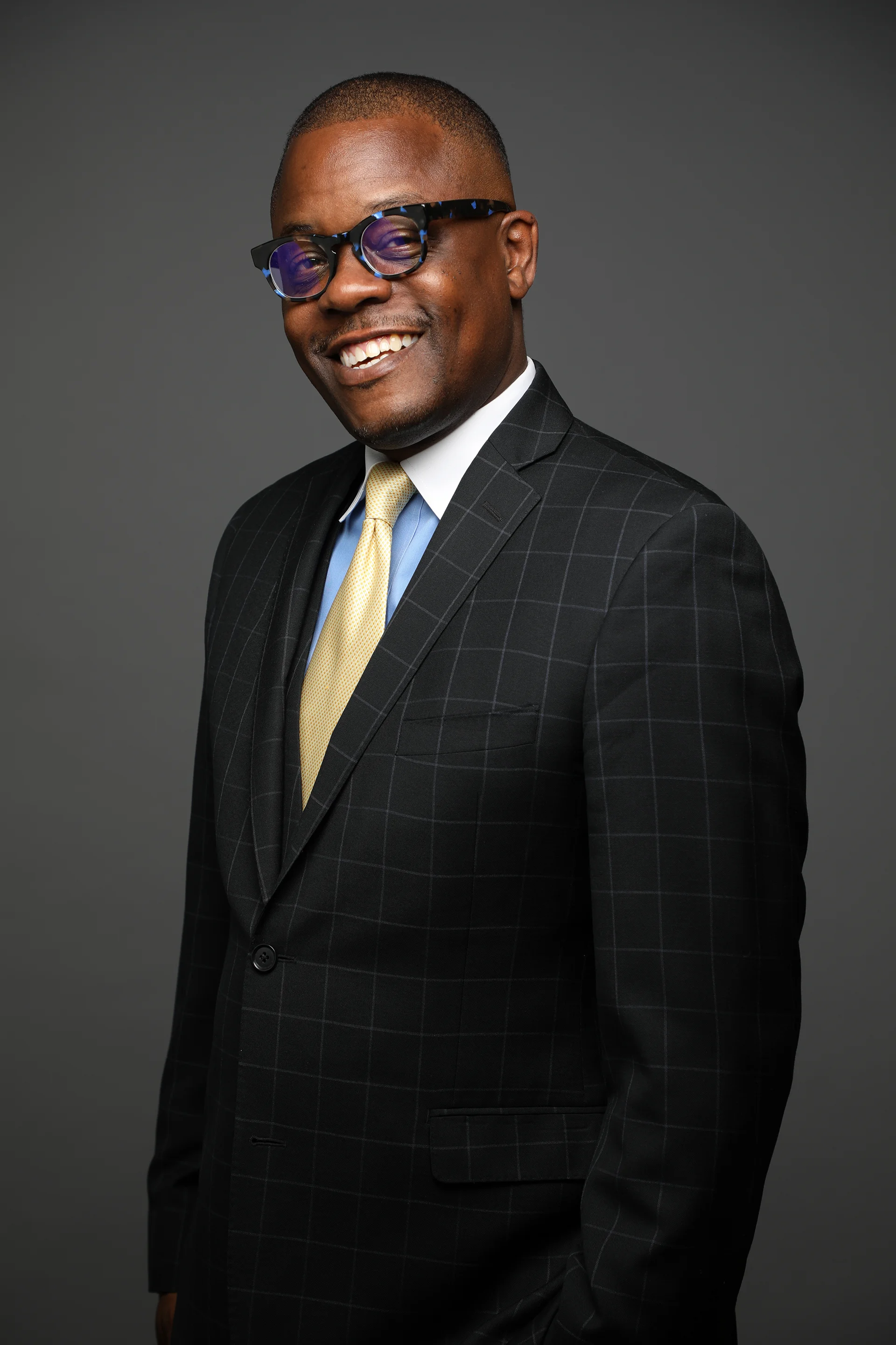 A smiling man wearing a black checkered suit, white shirt, yellow tie, and tortoiseshell glasses against a gray background.