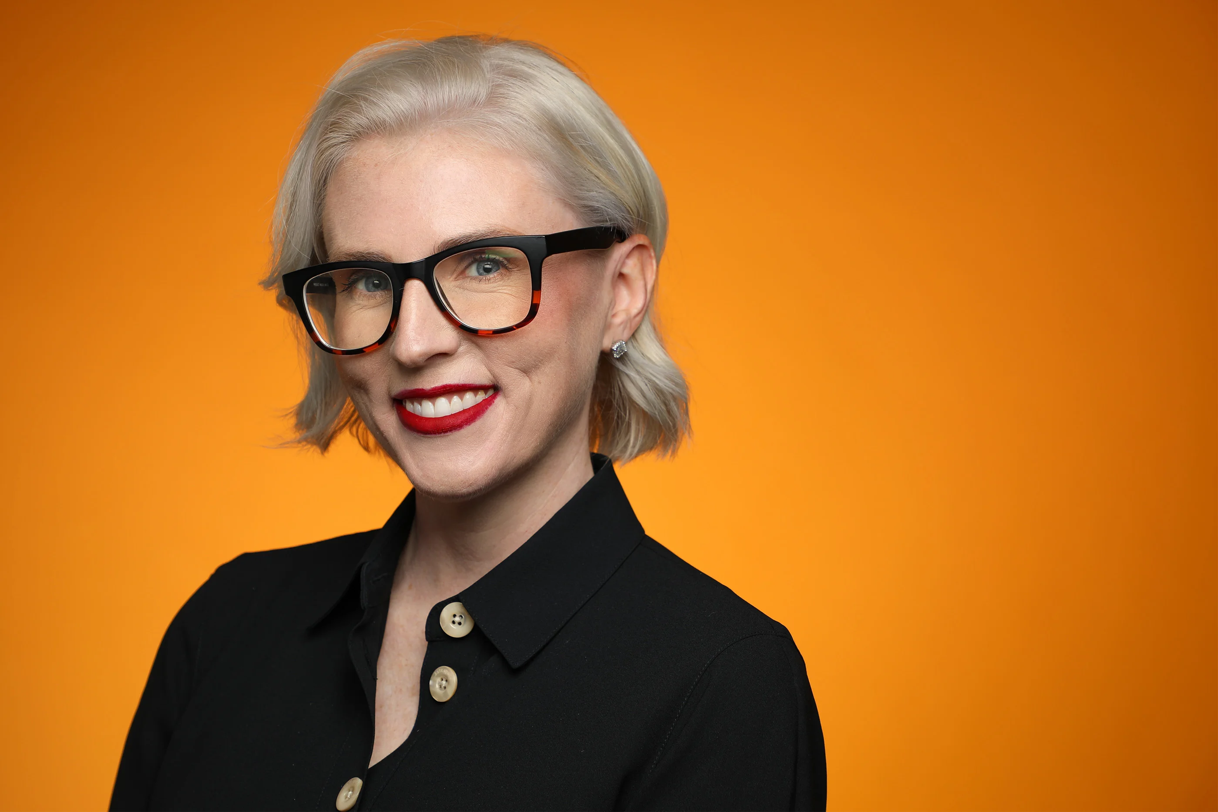 A woman with short blonde hair smiling, wearing black glasses with tortoise shell accents, red lipstick, a black button-up shirt with pearl buttons, and earrings, against an orange background.