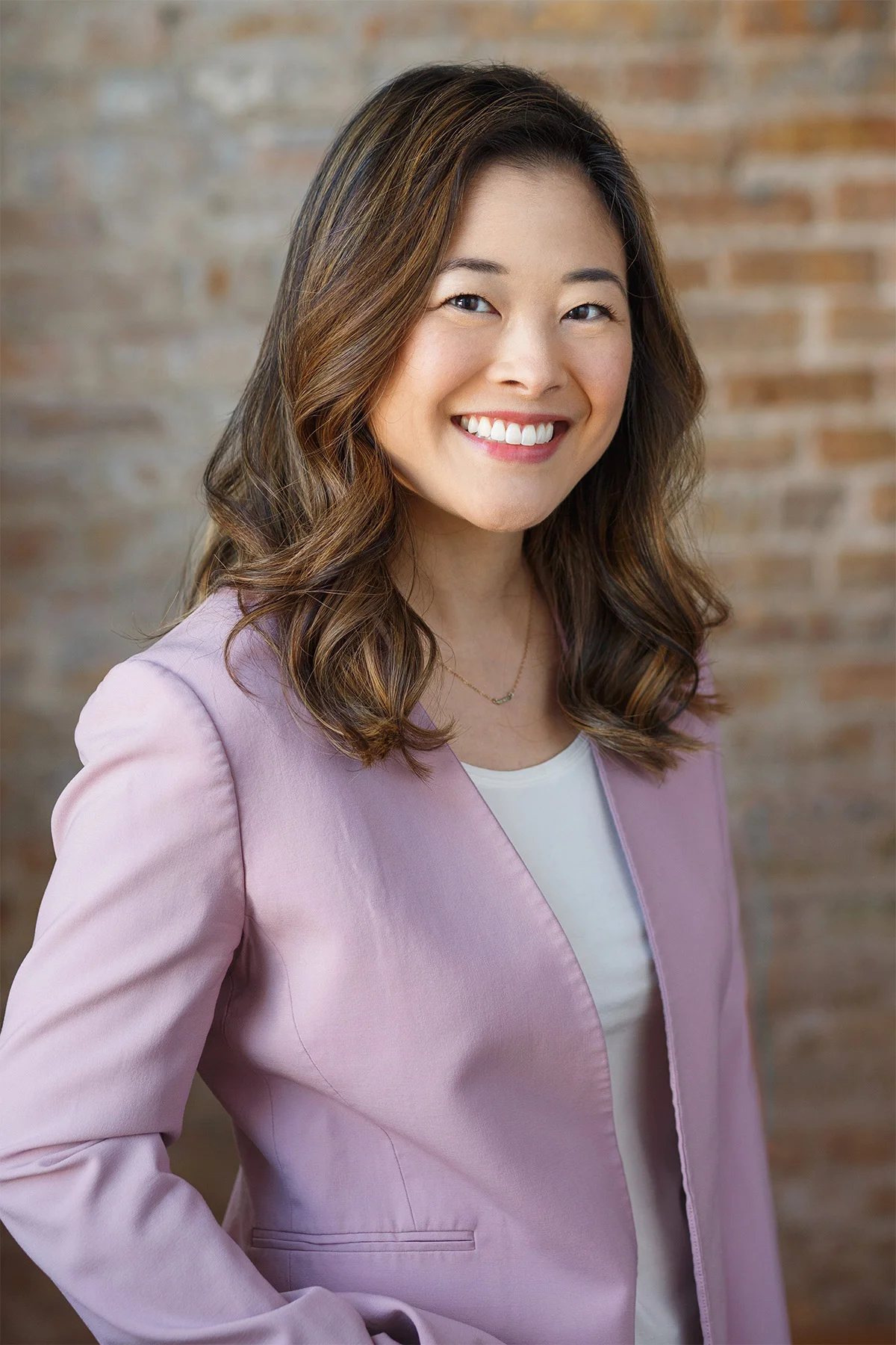 A woman with long, wavy brown hair and blue eyes smiling, wearing a light pink blazer and a white top, standing against a blurred brick wall background.