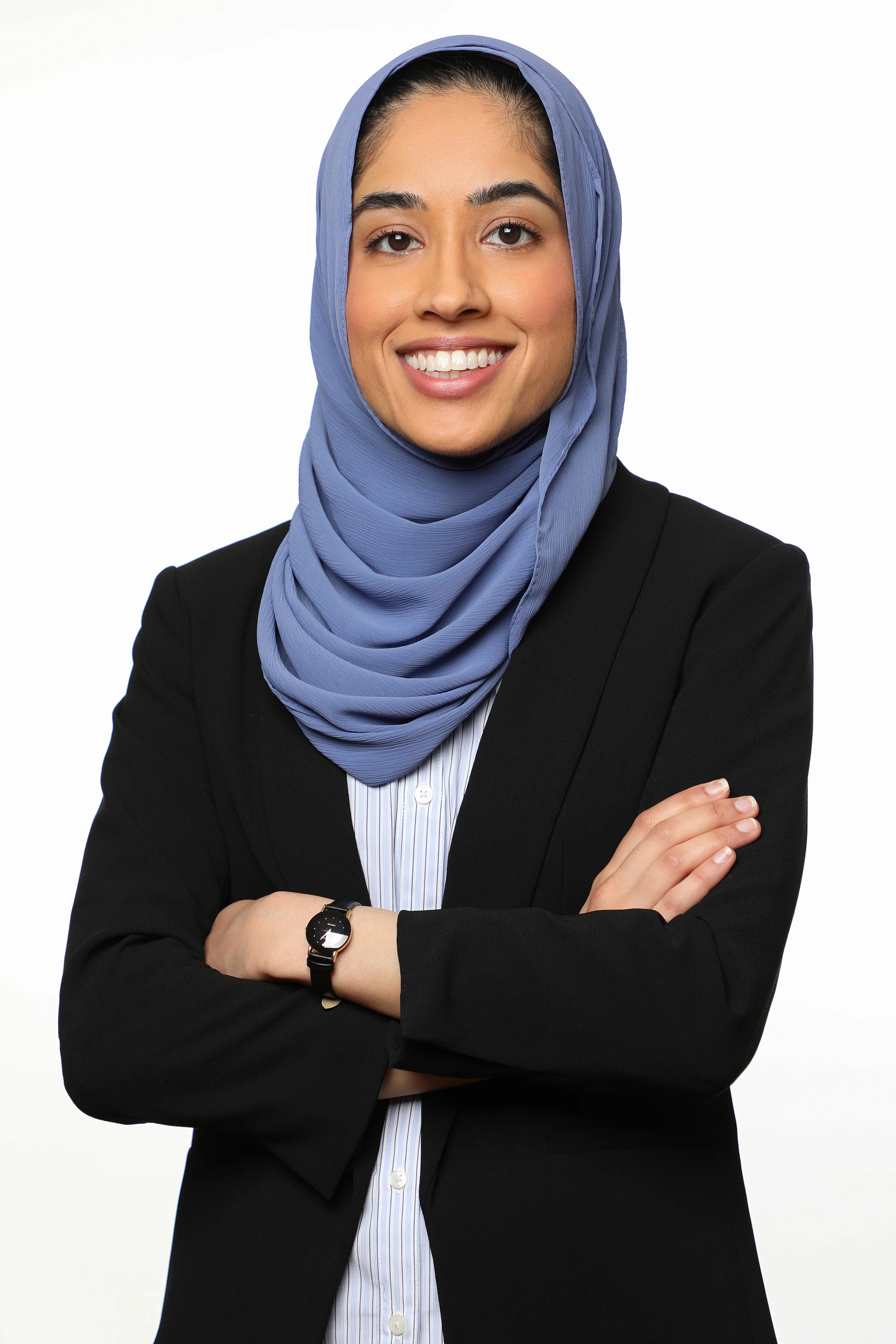 Professional woman wearing a black blazer, a light blue striped shirt, and a matching blue hijab, smiling with crossed arms, against a white background.