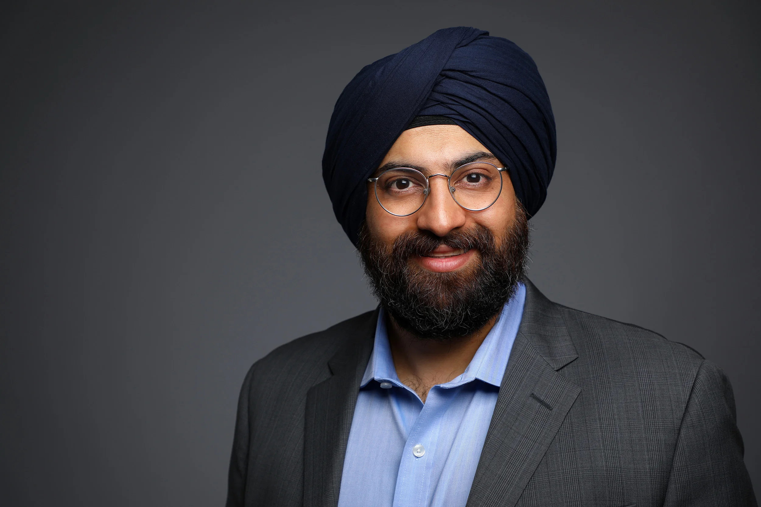 Portrait of a man wearing glasses and a turban, dressed in a suit and blue shirt, smiling against a gray background.