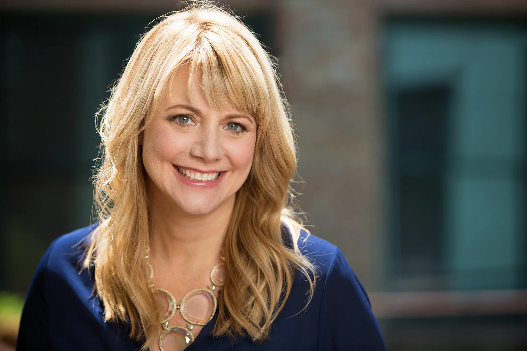 Close-up of a smiling blonde woman wearing a blue blazer and a necklace, standing outdoors with a blurred background.