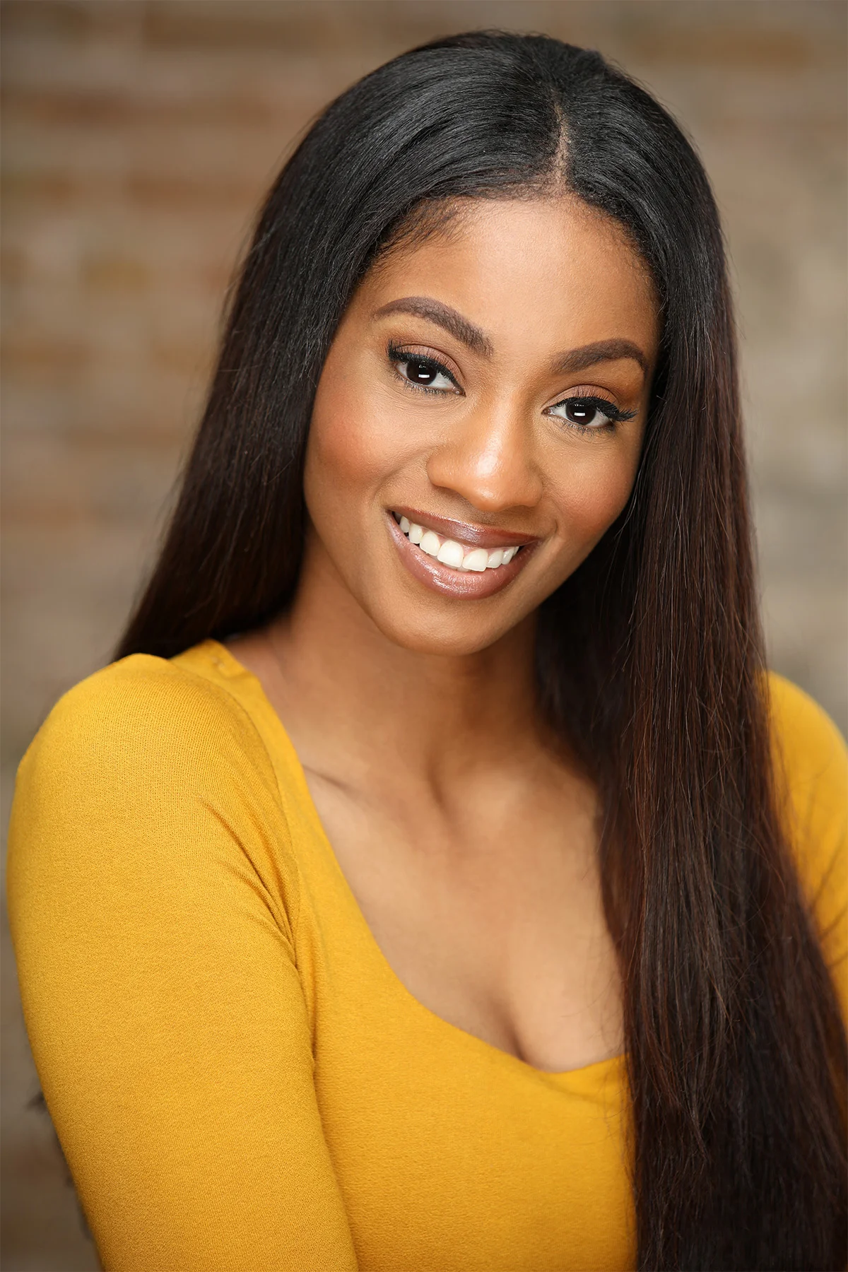 A smiling woman with long dark hair and brown skin wearing a yellow top, posing with a blurred brick wall background.
