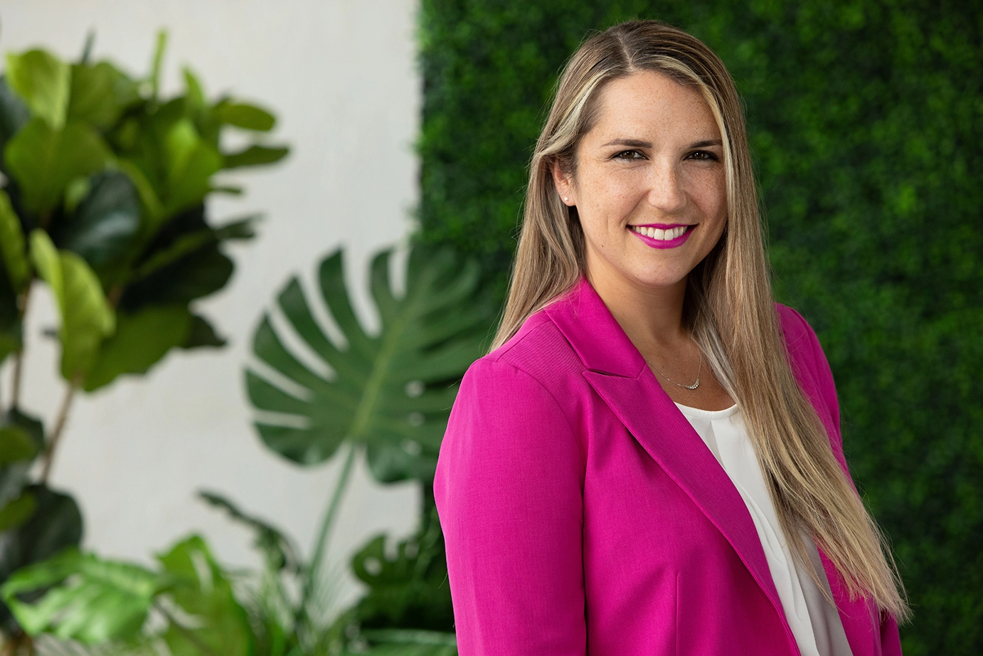 A woman with long blonde hair wearing a bright pink blazer and white top, smiling, standing in front of a lush green plant and blurred greenery background.