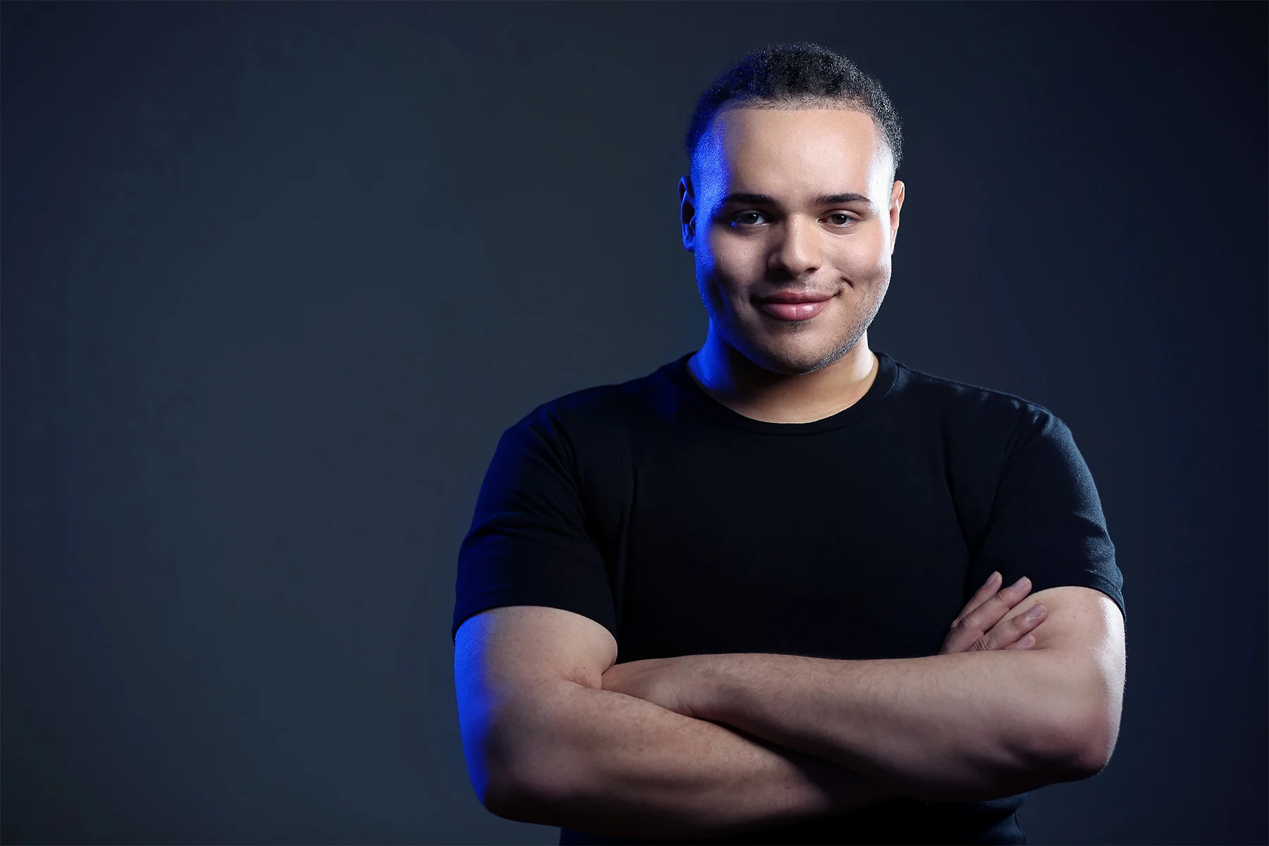 A young man with short hair and a slight smile, crossing his arms and wearing a black t-shirt, standing against a dark background with blue lighting on one side.