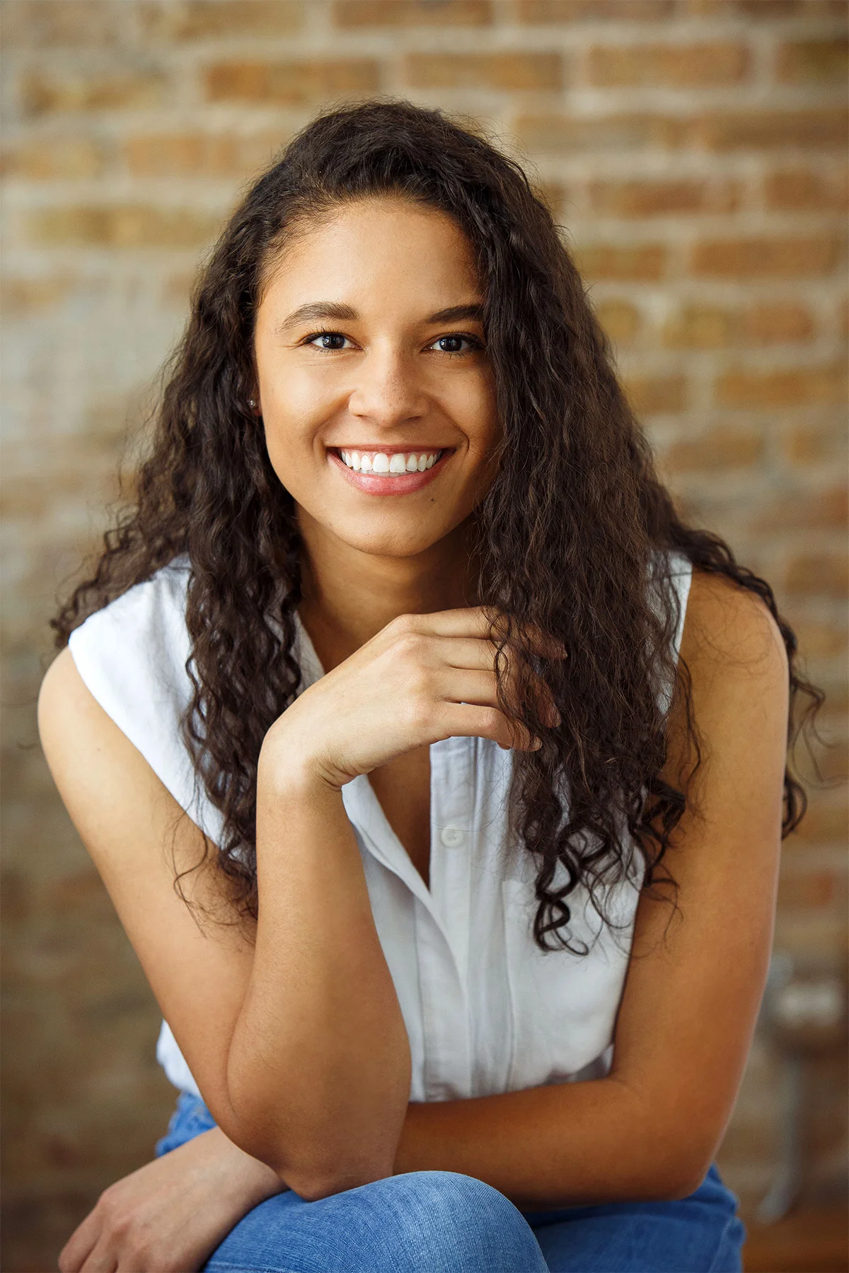 A young woman with long curly dark hair, smiling and sitting with her hand resting on her chin in front of a brick wall background.