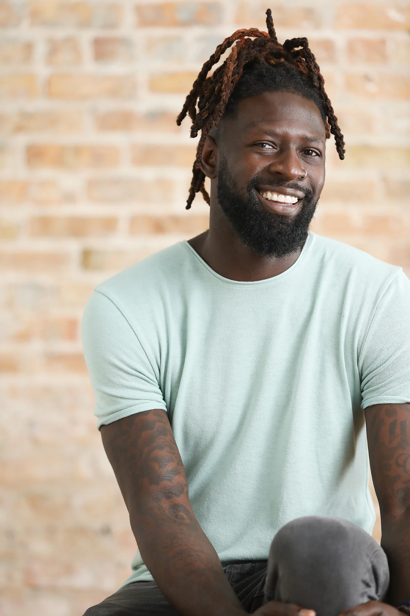 A smiling man with dreadlocks pulled up, a nose ring, and a beard, wearing a light green t-shirt, sitting against a brick wall background.