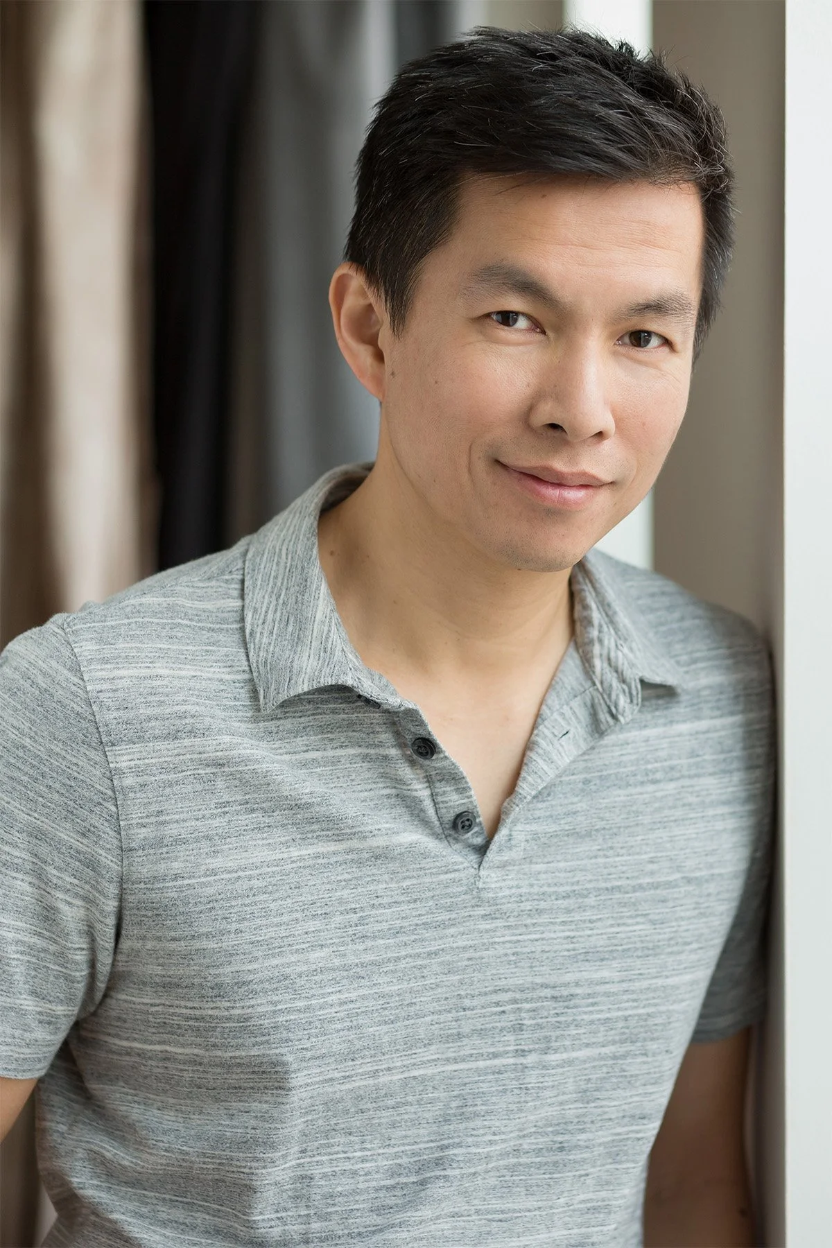 A man with short black hair wearing a light gray polo shirt standing near a window, smiling softly.