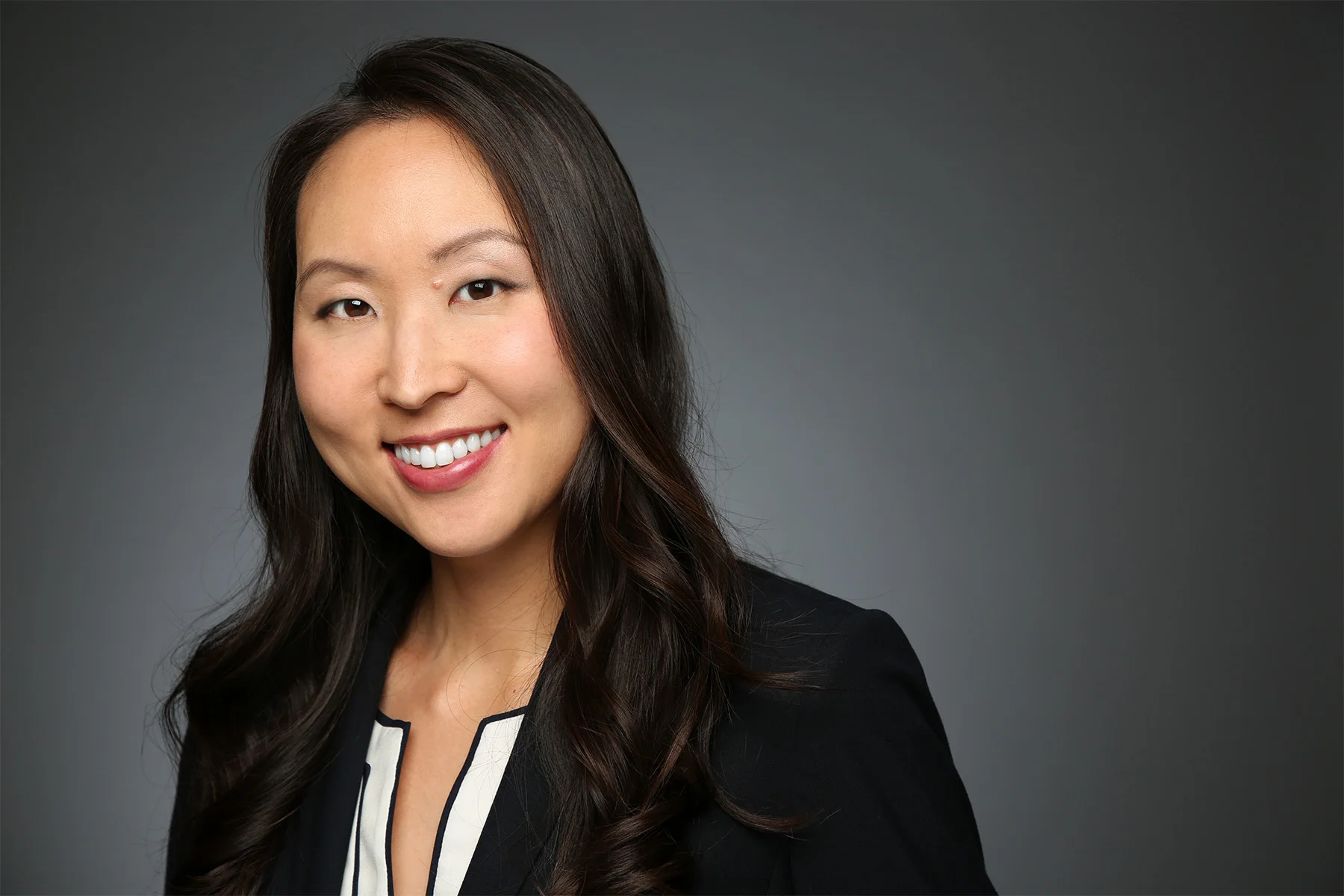 Professional headshot of an Asian woman with long dark hair, smiling, wearing a black blazer and a white shirt with black trim, against a dark gray background.