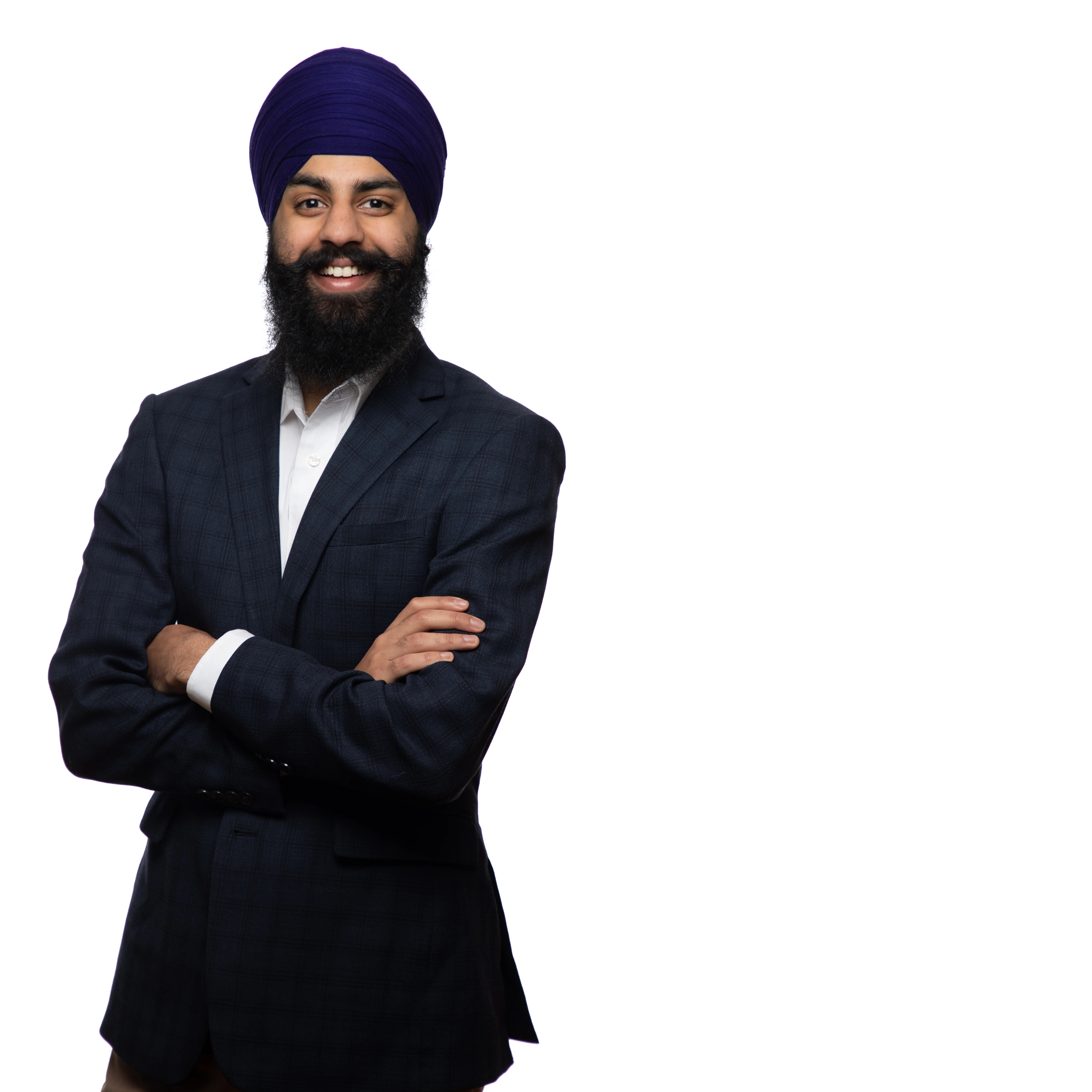 A smiling man wearing a blue turban, dark checkered suit, and white shirt, standing with arms crossed against a white background.