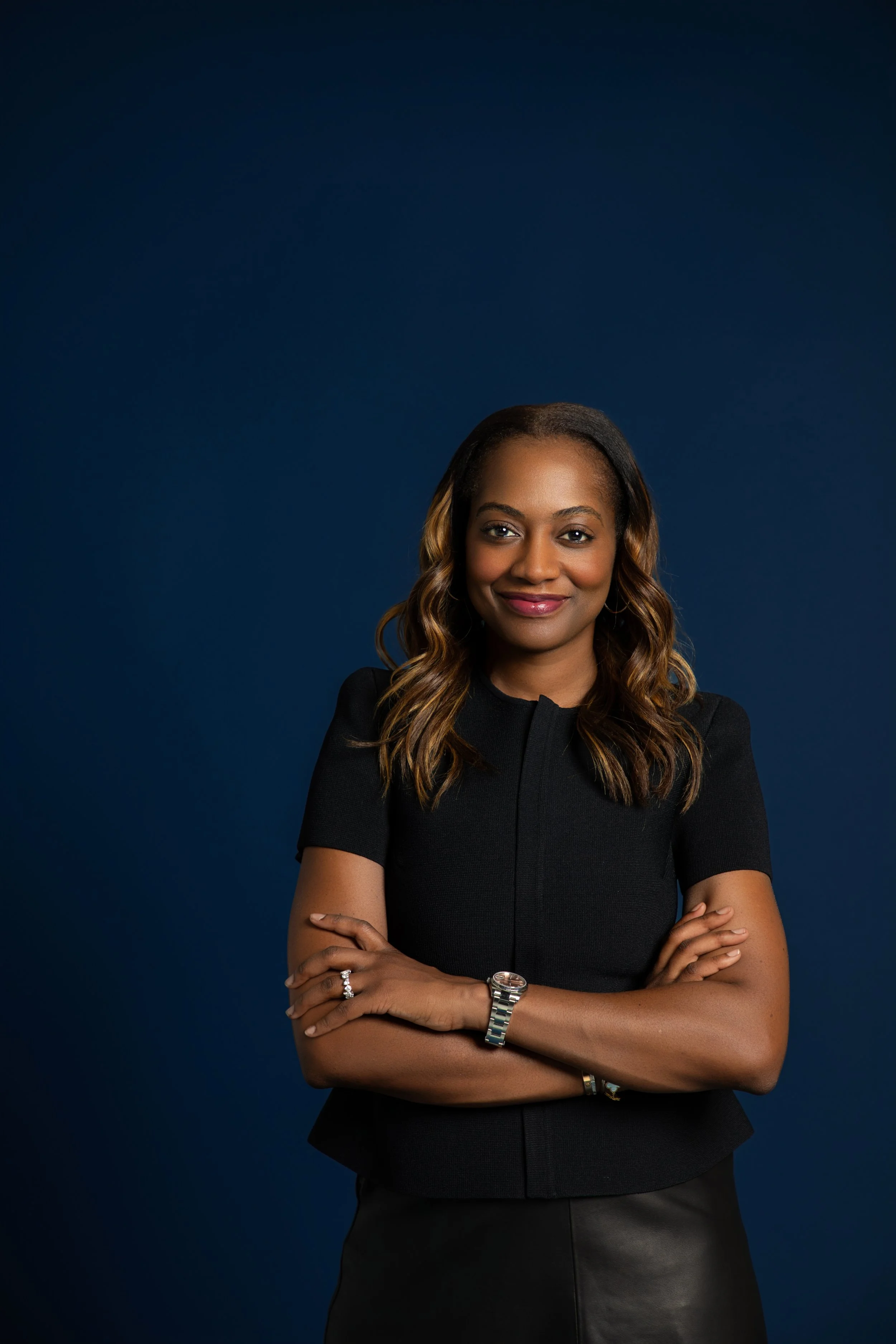 A professional woman with long wavy hair, wearing a black outfit, standing against a dark blue background with her arms crossed, smiling confidently.