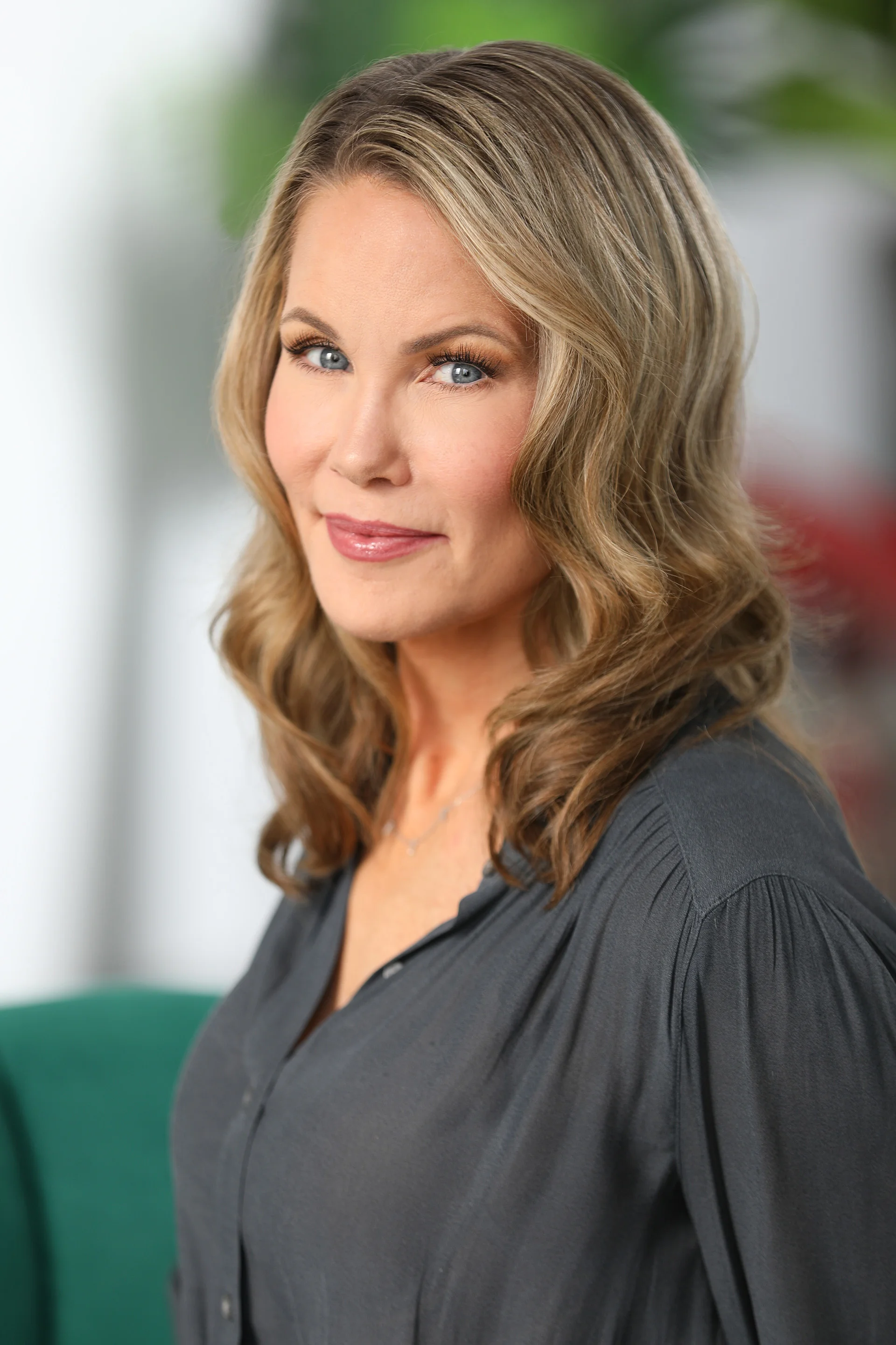 A woman with light skin, blond wavy hair, blue eyes, and makeup, wearing a dark gray top, smiling softly, with a blurred indoor background.