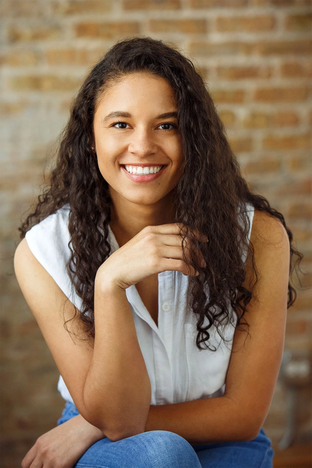 A young woman with long curly brown hair, smiling, wearing a white sleeveless blouse and blue jeans, sitting in front of a brick wall.