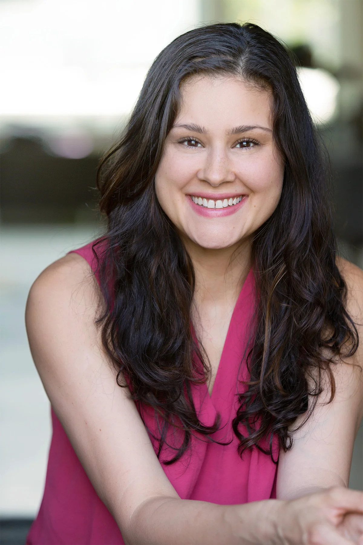 A young woman with long curly dark hair, smiling and wearing a pink sleeveless top, sitting indoors with a blurred light background.
