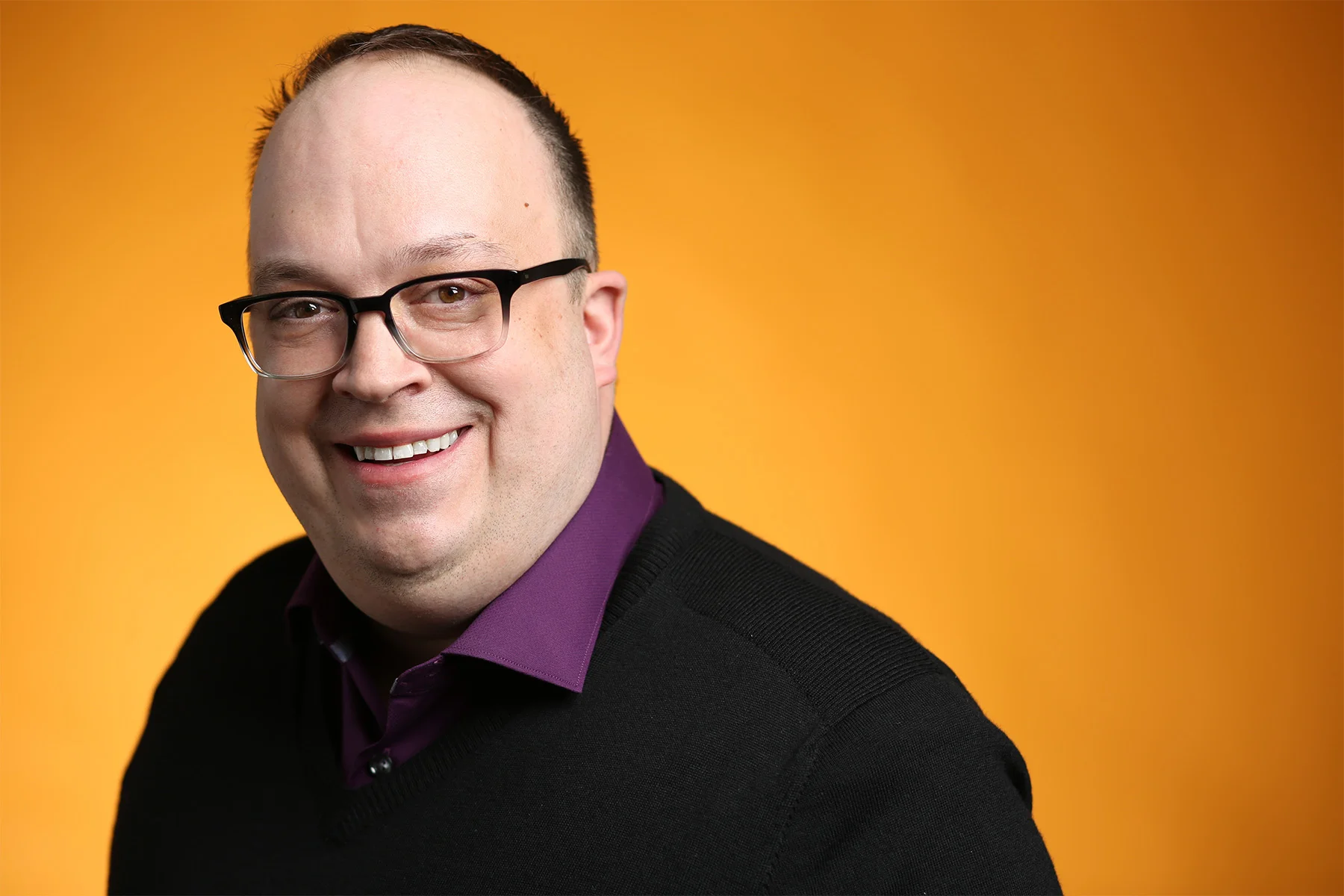 A smiling man with glasses, wearing a black sweater over a purple shirt, against an orange background.