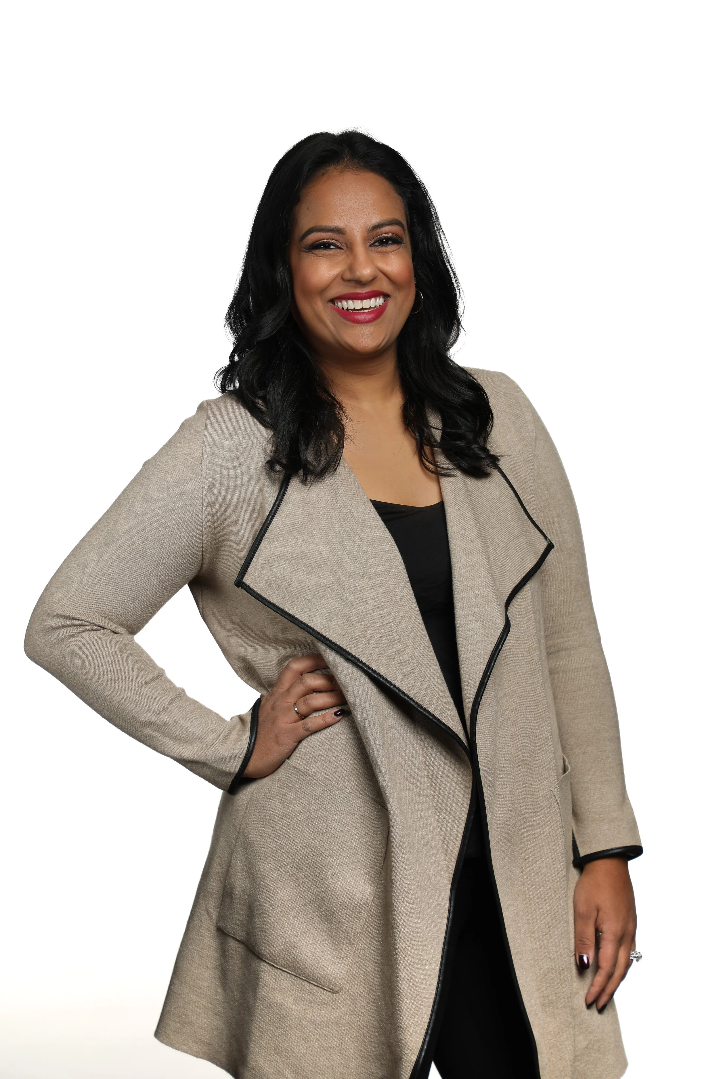 A woman with medium skin tone, black wavy hair, dressed in a beige coat with black trim, standing confidently with her hand on her hip against a white background.