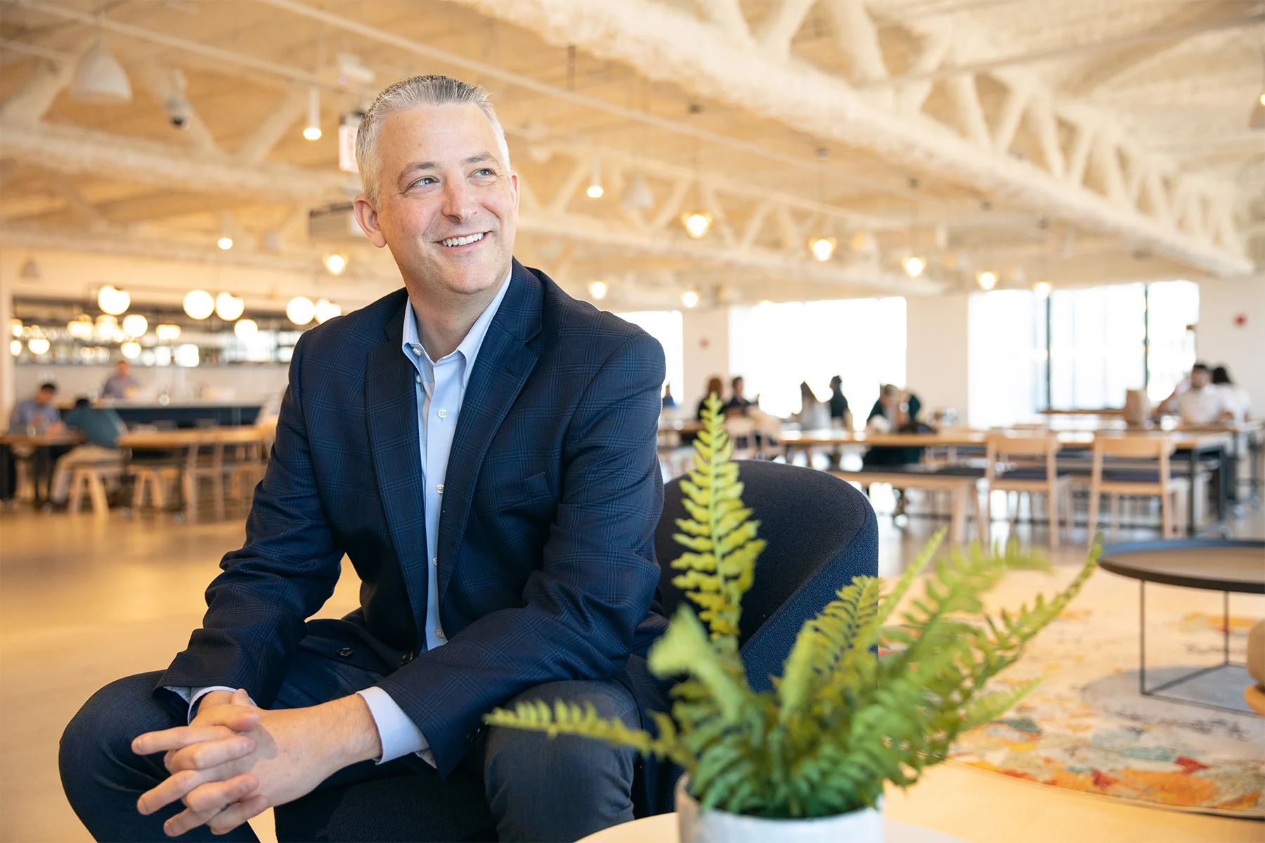 A smiling man in a navy blue suit sitting in a modern, well-lit office or coworking space with wooden beams and large windows in the background, with green plants in the foreground.