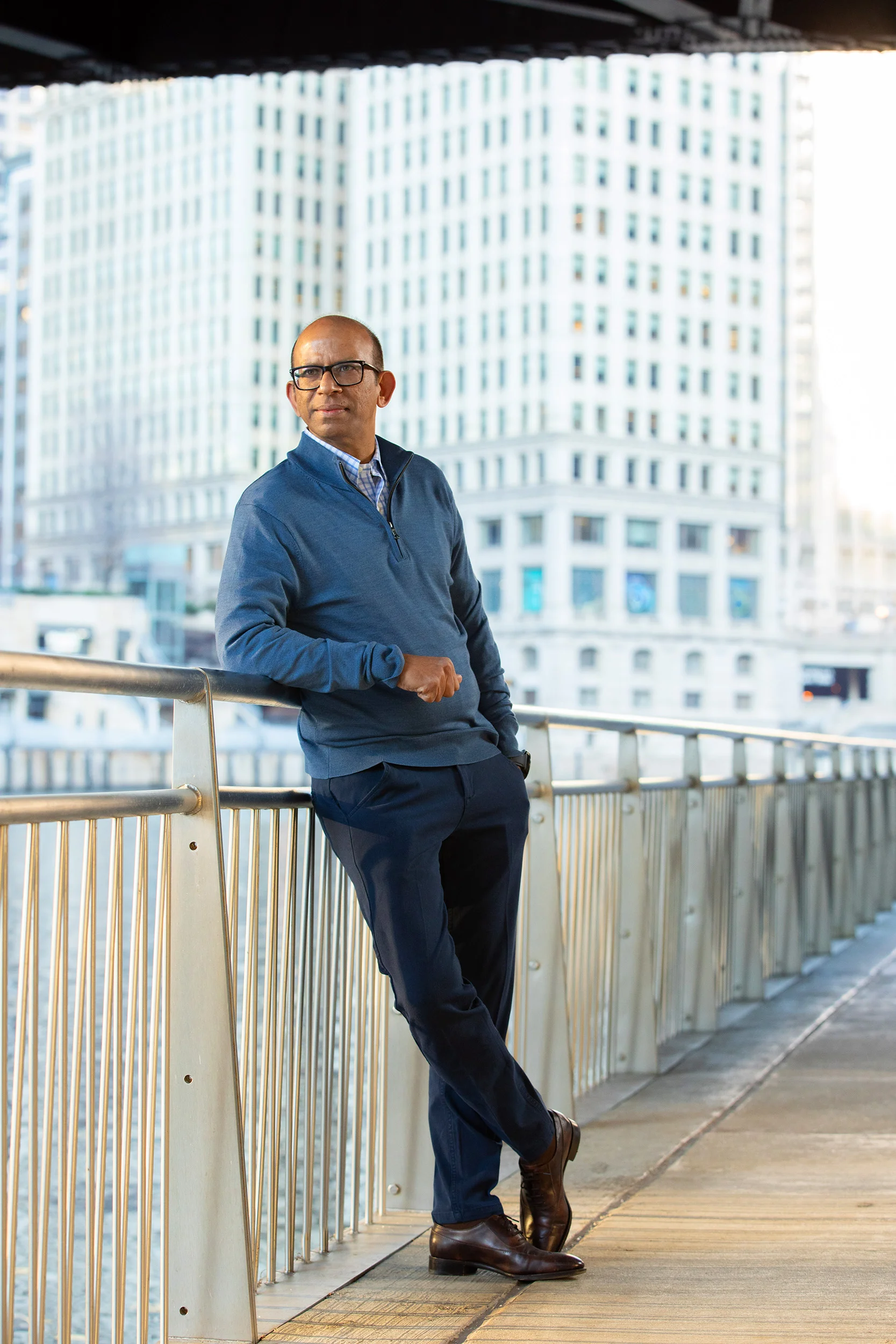 A man wearing glasses, a blue quarter-zip sweater, and dark pants, stands on a balcony with a cityscape of tall, modern buildings in the background. He is leaning against a metal railing with one hand and has his other hand in his pocket, looking sli