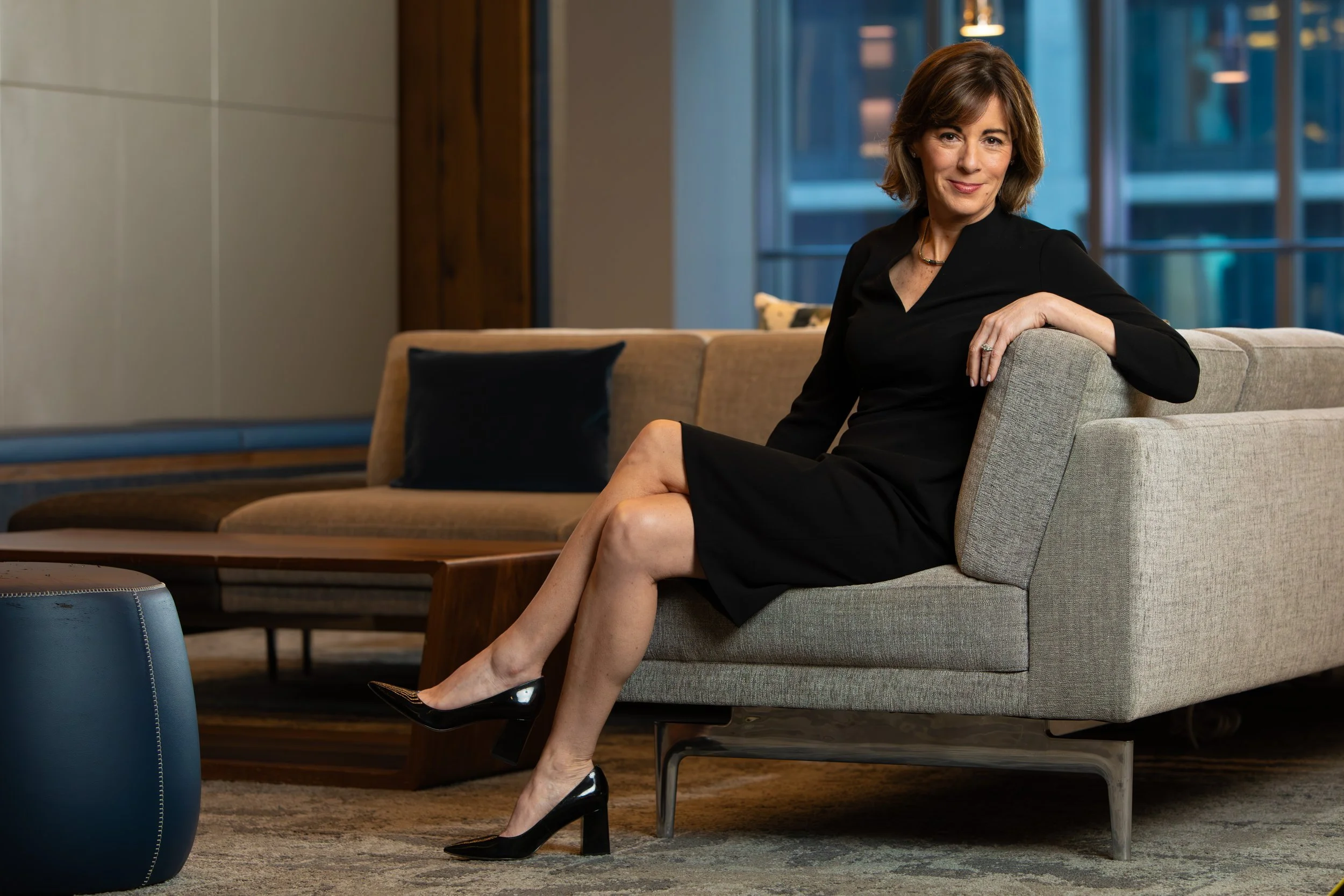 A woman with short brown hair, wearing a black dress and black high heels, sitting on a light gray sofa in a modern lounge area with large windows, wooden accents, and a coffee table.