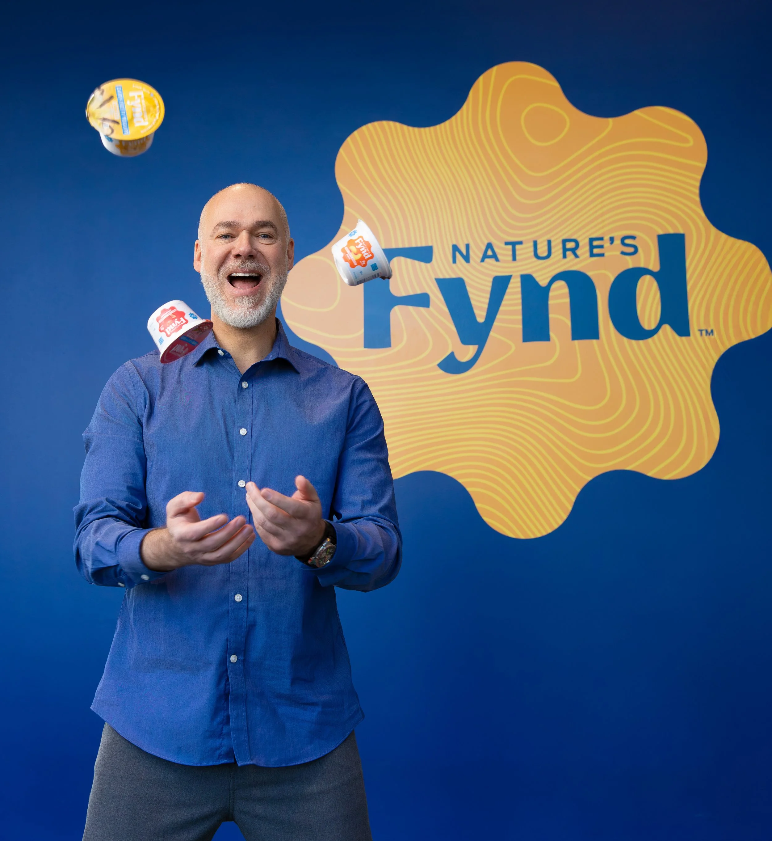 A man with gray hair and beard wearing a blue button-up shirt, smiling, with yogurt containers flying around him against a blue background with the logo 'Nature's FYND' and a yellow abstract shape.
