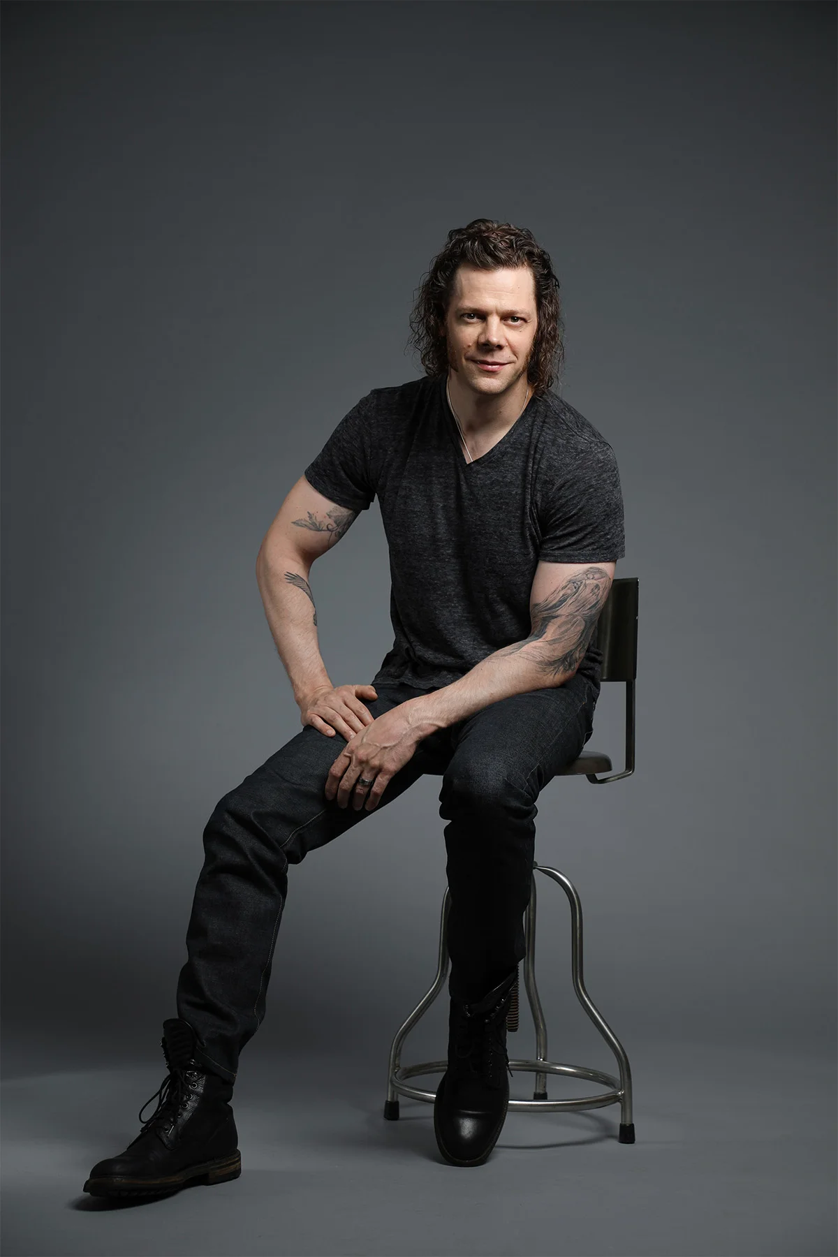 A man with curly dark hair and tattoos, dressed in black, sitting on a stool in front of a grey background. He is looking at the camera with a slight smile.