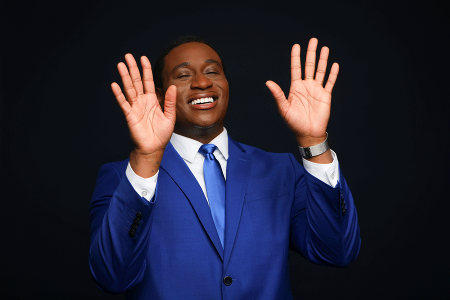 A man in a blue suit smiling with eyes closed, raising both hands near his face against a dark background.