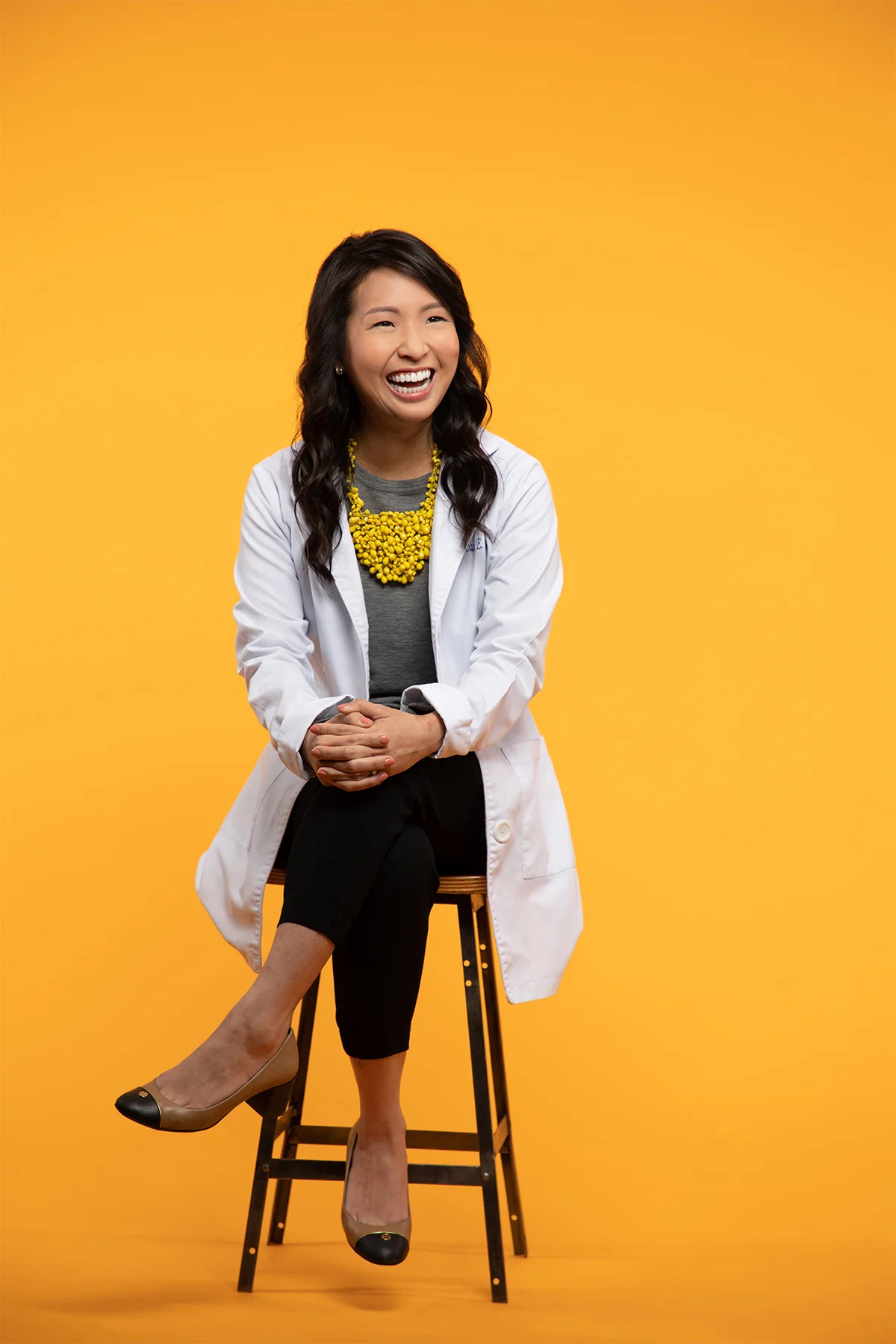 A smiling woman sitting on a stool in front of a yellow background. She has long dark hair, is wearing a white lab coat, a yellow beaded necklace, gray shirt, black pants, and beige high heels.